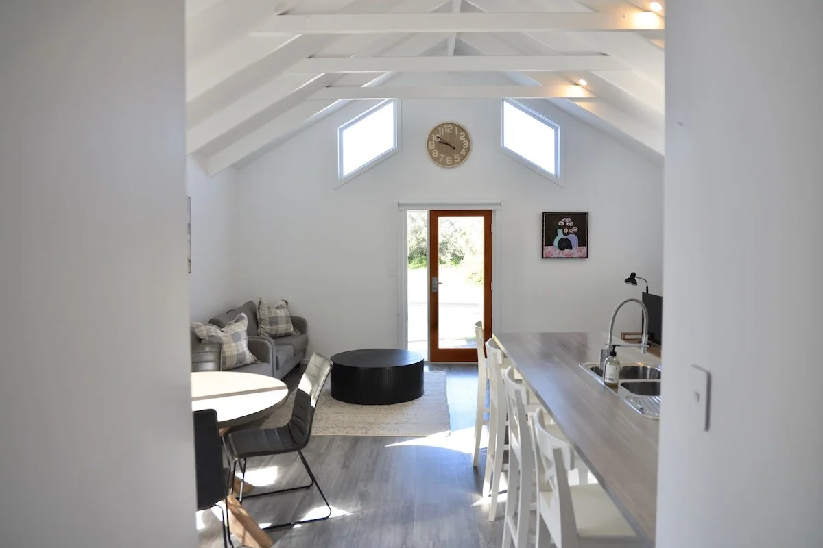 Interior view of a bright living room and kitchen area with vaulted ceiling, two skylights, a wall clock, a gray sofa, a dining table with black chairs, a bar counter with stools, and a door leading outside.