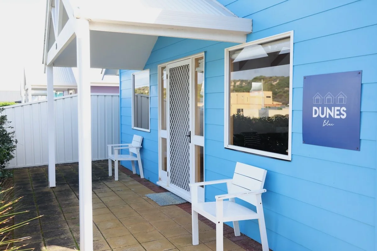 Blue beachfront accommodation with two white chairs, a door with a security screen, windows, and a sign that reads 'Dunes Blu' on the exterior wall.