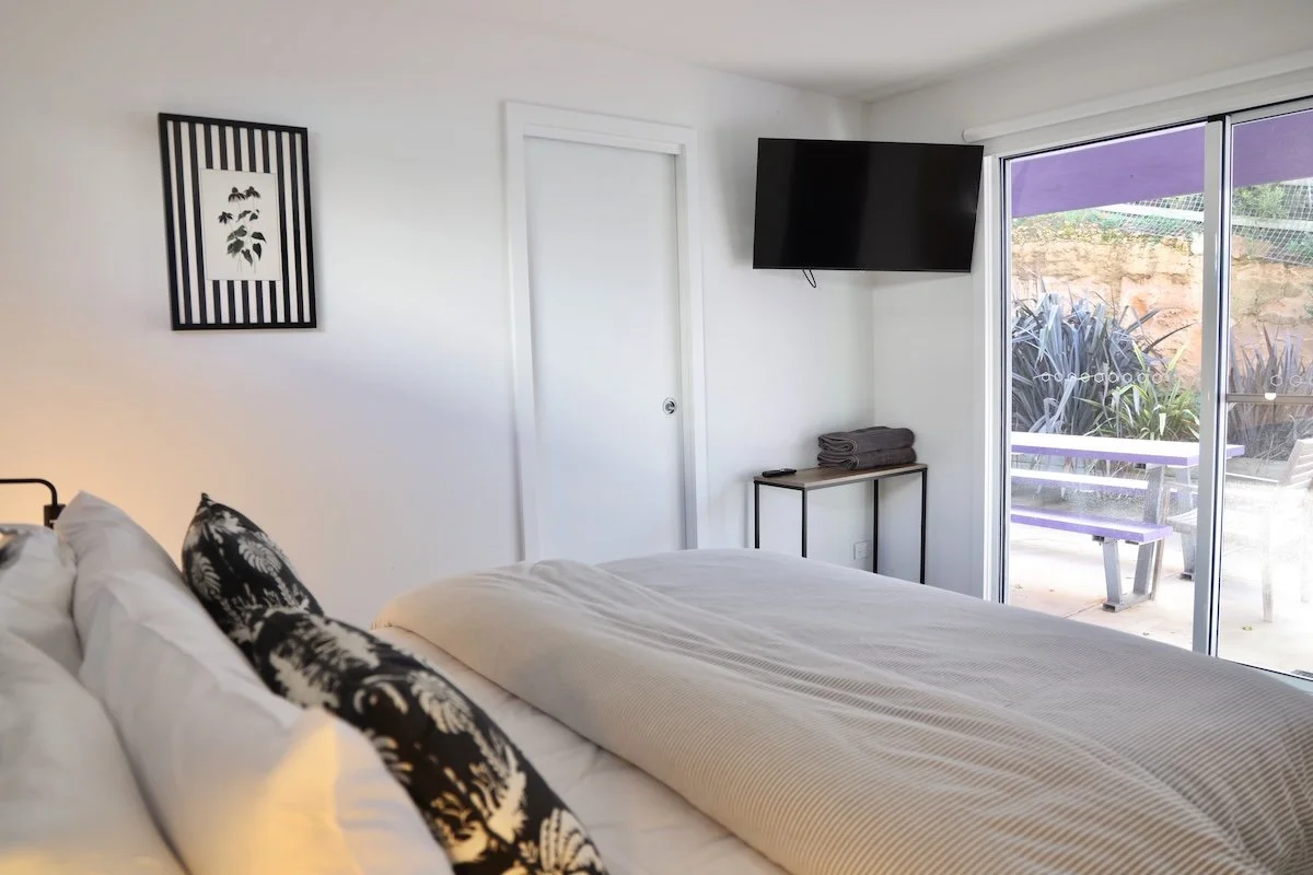 A bedroom with a bed, black and white pillows, a wall-mounted TV, a sliding glass door to an outdoor patio, and a framed black and white botanical artwork on the wall.