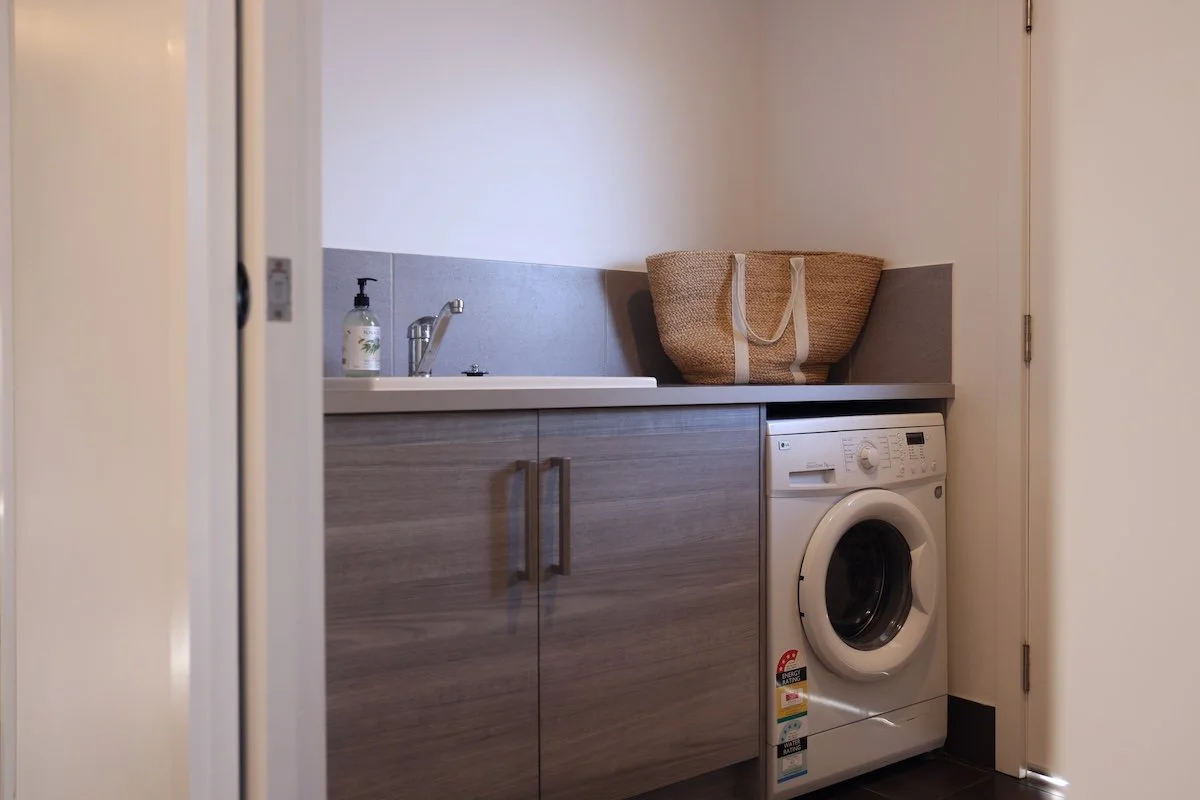 A laundry room with a washing machine under a countertop, a cabinet with handles, a soap dispenser, and a woven storage basket on the counter.