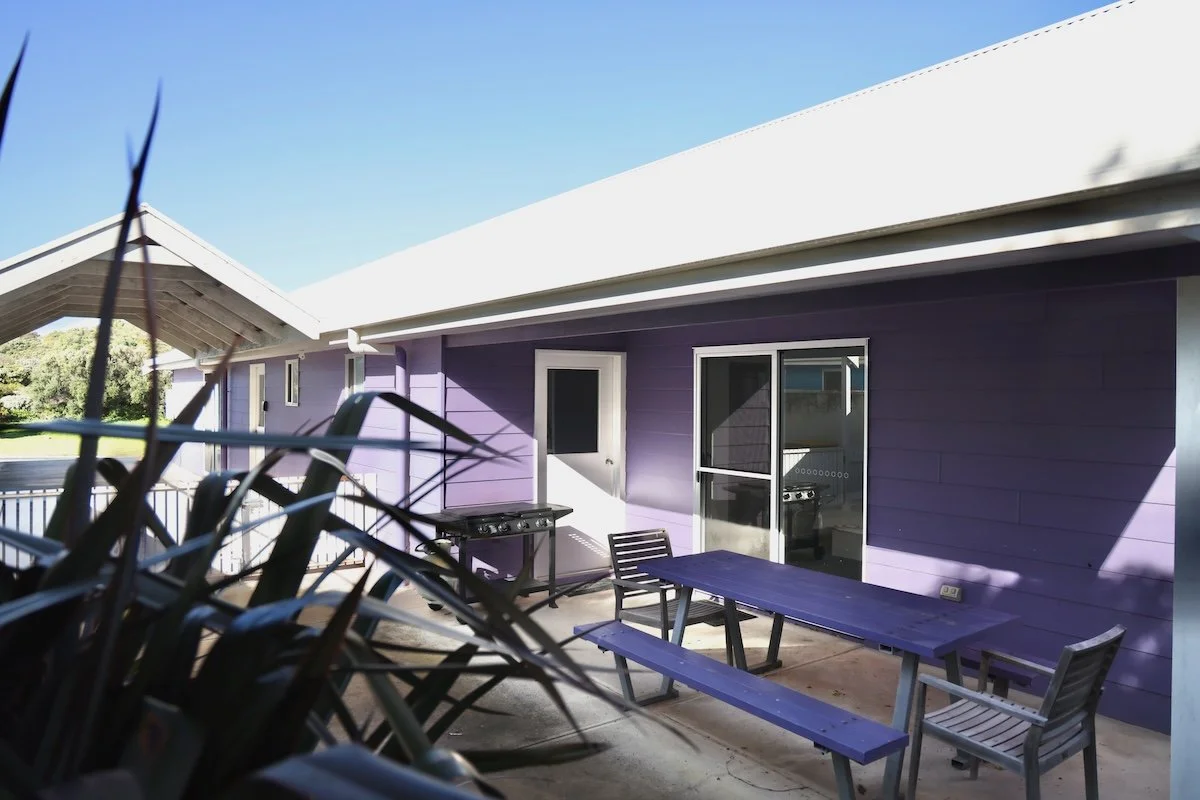Purple house with an outdoor patio area featuring a purple picnic table and benches, a grill, and chairs, with a clear blue sky overhead.