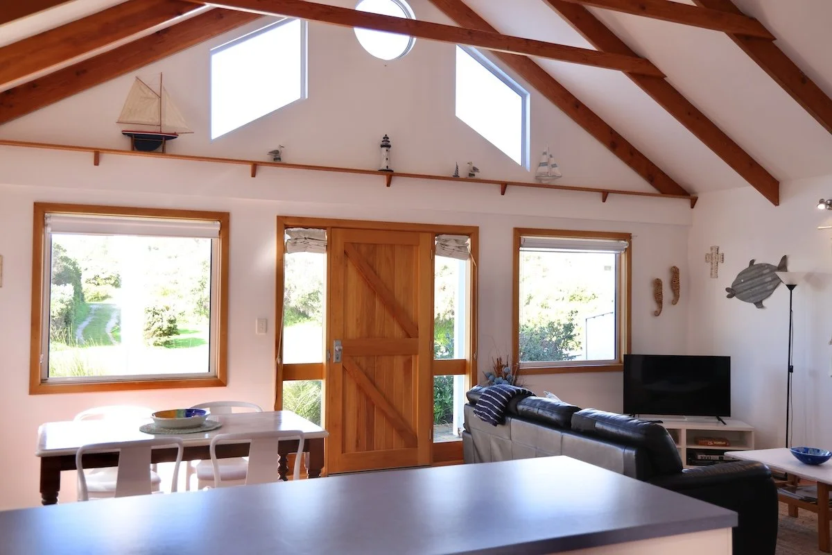 Living room with high vaulted ceiling and wooden beams, three windows, a wooden door, a dining table with chairs, a sofa, a TV, and nautical decor including model ships and fish wall art.
