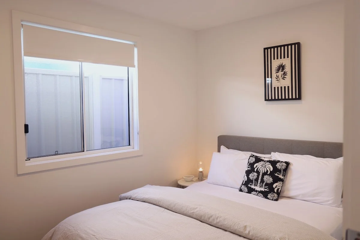 A neatly made bed with white sheets, pillows, and a black decorative pillow featuring a palm tree pattern in a bedroom with white walls. There is a window with a white blind on the left and a framed black and white wall art above the bed. A small nig
