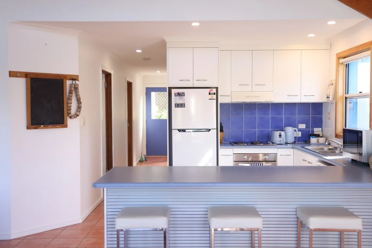 Kitchen with white cabinets, blue backsplash, stainless steel stove, refrigerator, microwave, and small kitchen island with three stools, and a doorway at the end of the hallway.