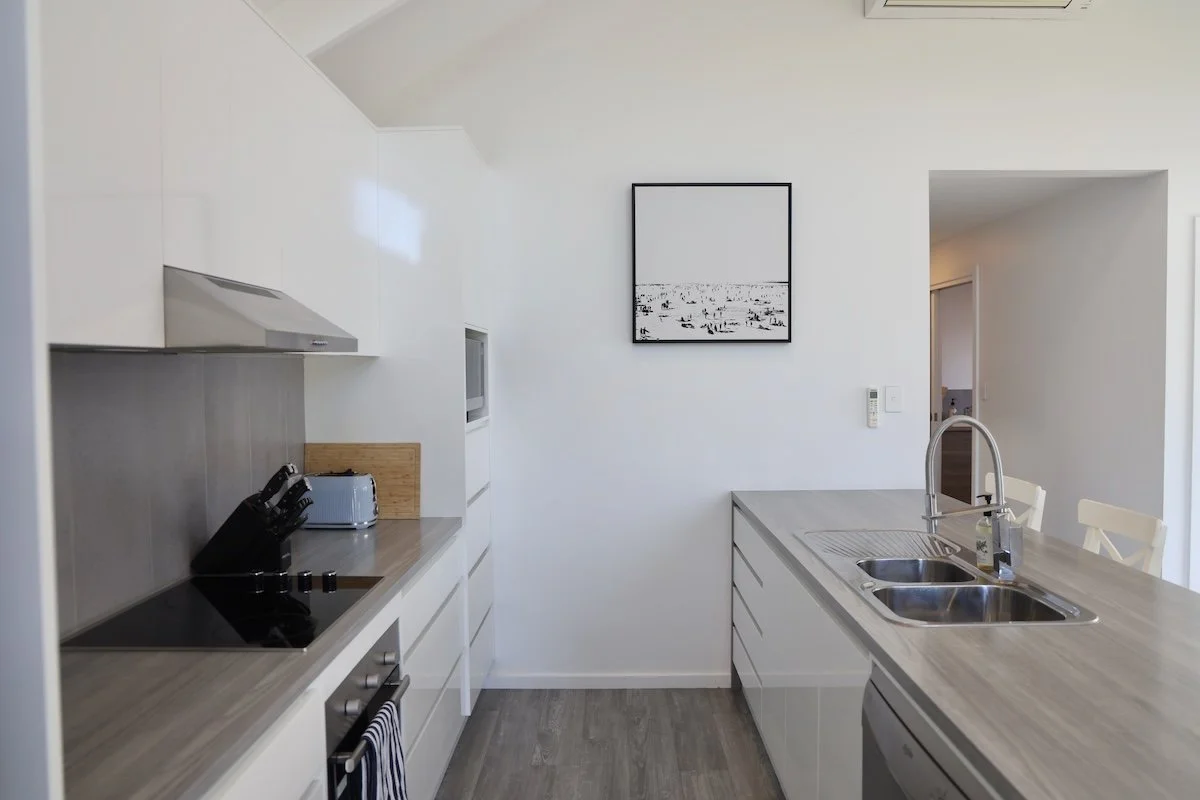 Modern kitchen with white cabinets, a gray countertop, a black cooktop, a microwave, a toaster, and a knife block. There is a black and white framed picture on the wall, a sink with a soap dispenser, and a bar stool.