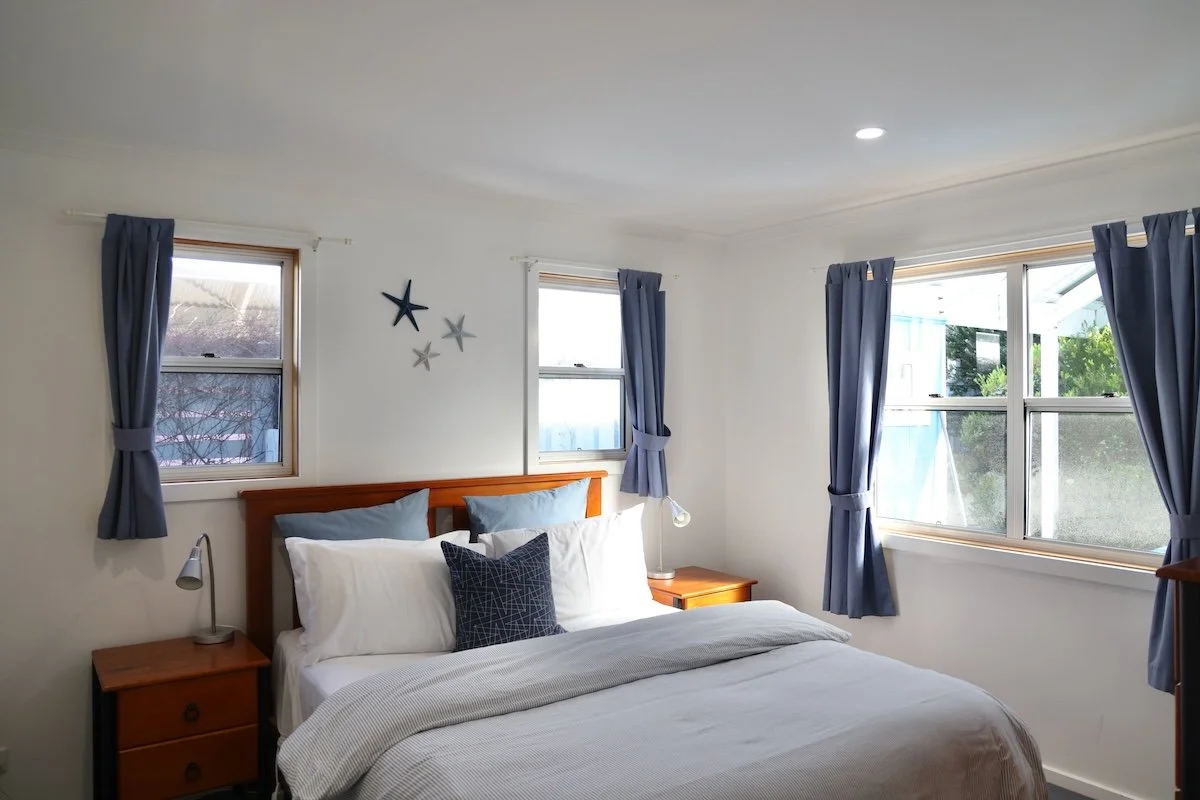 A neatly made bed with white bedding, blue pillows, and a brown wooden headboard in a bedroom with three windows, blue curtains, and wall decorations.