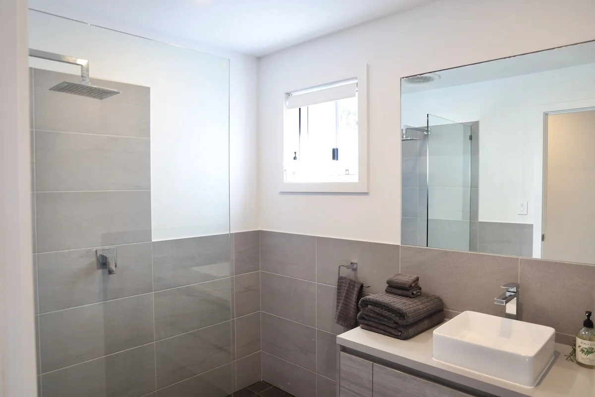 Modern bathroom with a walk-in shower, a large mirror, and a white rectangular sink on a grey cabinet, with folded dark towels and a soap dispenser on the countertop, and a small window with blinds.