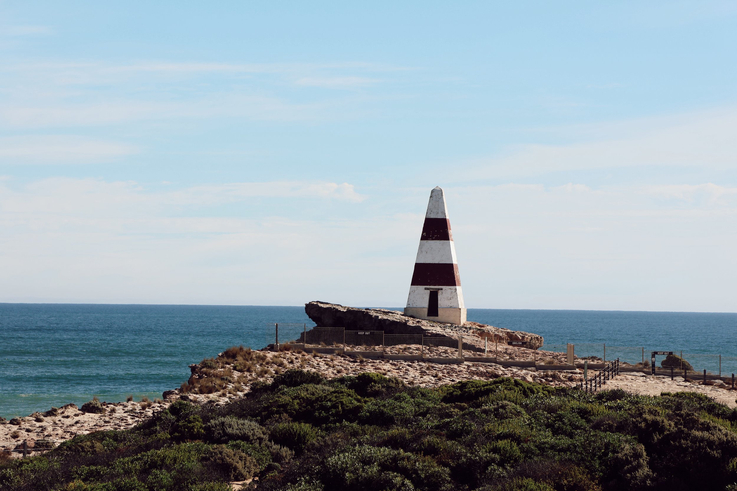 A lighthouse shaped like a striped cone with red and white bands, standing on a rocky coastline near the ocean under a partly cloudy sky.