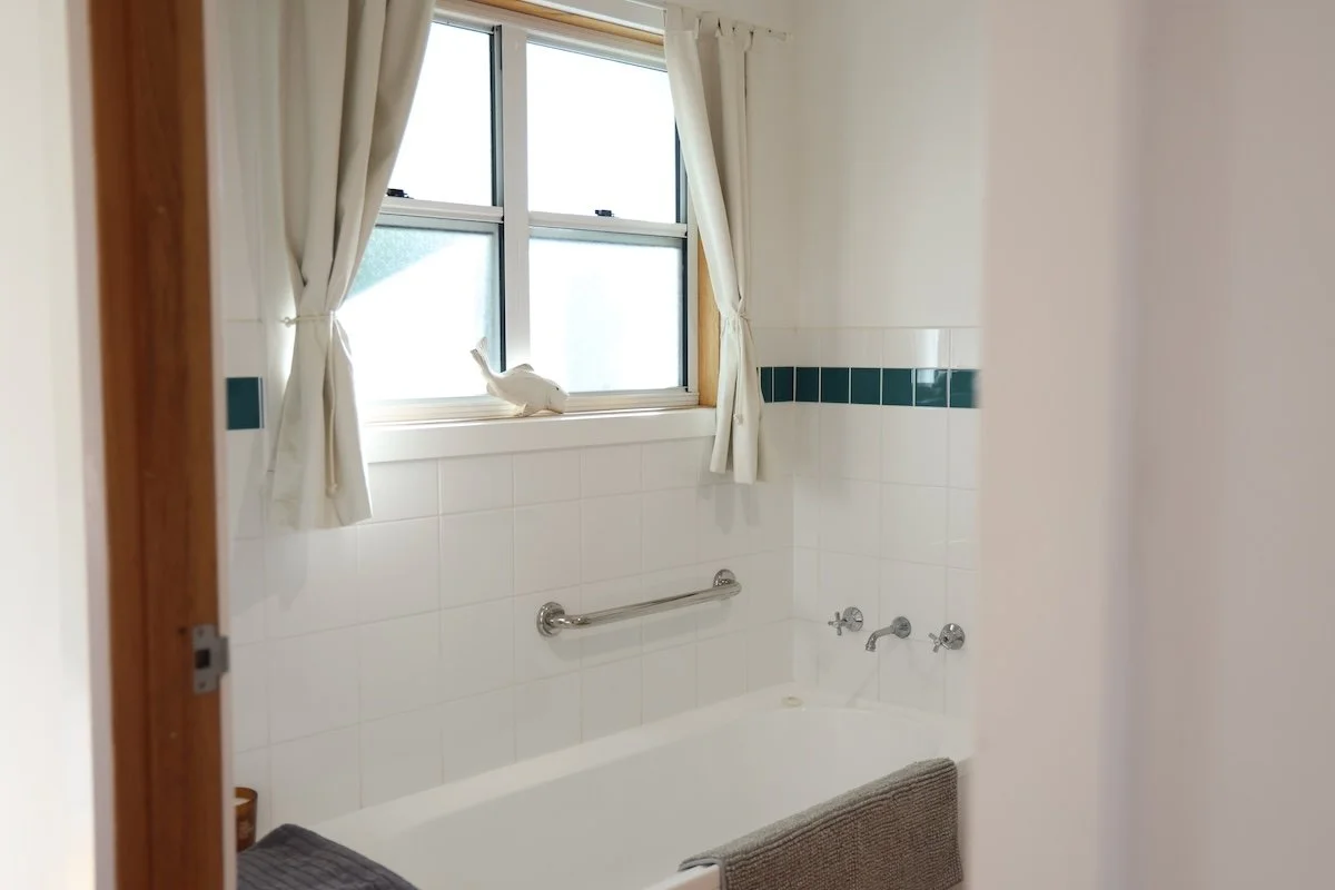 Bathroom with white tiles, a bathtub with a gray towel, a window with cream curtains, and a small decorative duck on the windowsill.