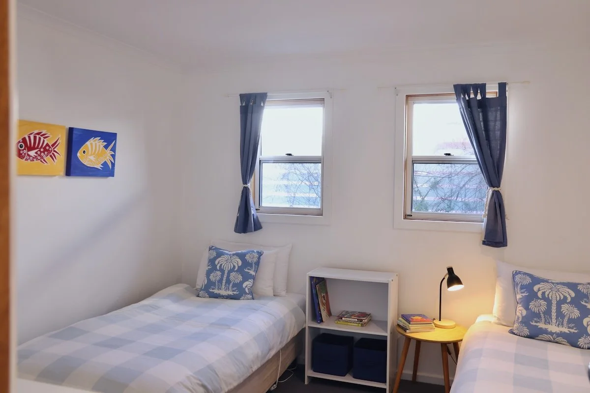 A bedroom with two twin beds, each with a pillow decorated with palm trees. There are two windows with blue curtains, a small white bookshelf with books and two storage bins, a wooden side table with a lamp and books, and two colorful fish paintings 