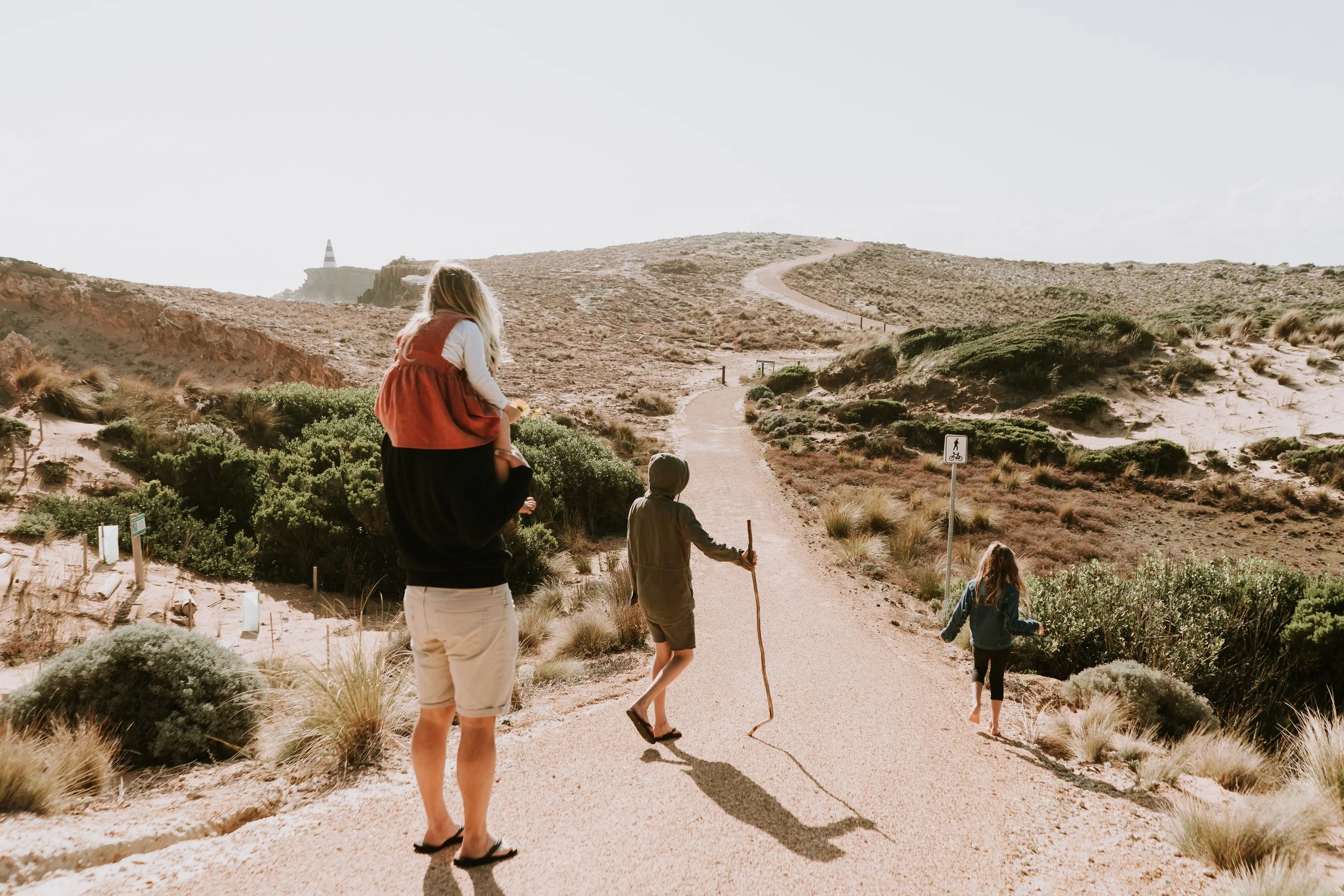 A group of four people hiking on a sandy trail in a desert landscape with low shrubs and sand dunes. One person is wearing a red vest, another is using a walking stick, and two children are with them. The trail is marked with a sign indicating a bicy