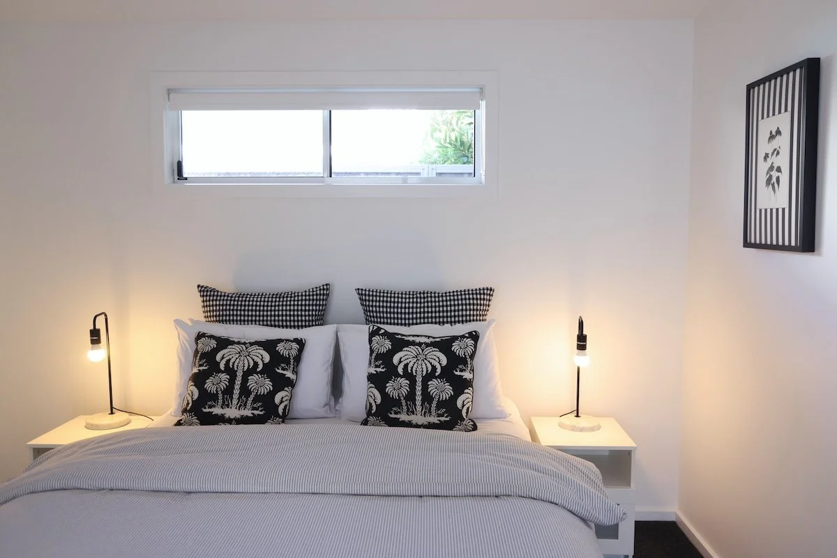 A bedroom with a white bed, black and white pillows, two bedside tables with modern lamps, a small horizontal window above the bed, and a framed black and white art piece on the wall.