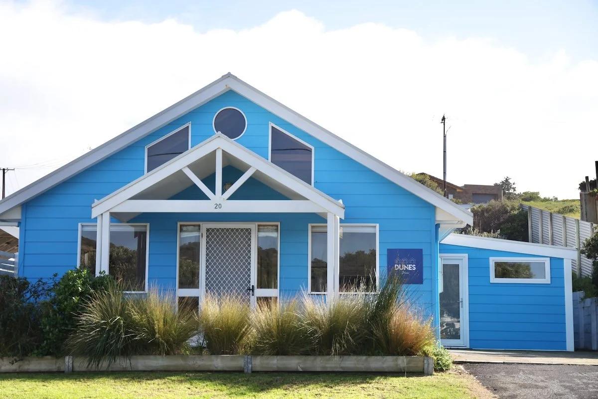 A bright blue beach house with white trim and a triangular roof, featuring a front porch with a white pergola, surrounded by grass and plants, with a sign that says "Dunes".