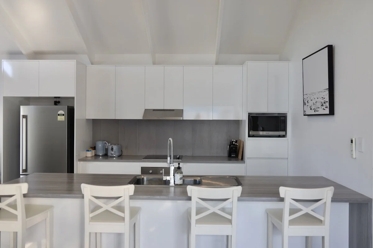 Modern white kitchen with gray countertops, stainless steel appliances, and four white chairs at a breakfast bar.
