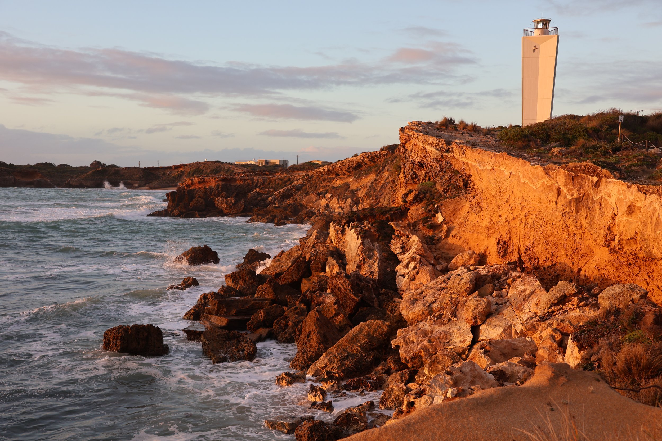 Coastline with rocky cliffs, ocean waves, and a lighthouse at sunset.