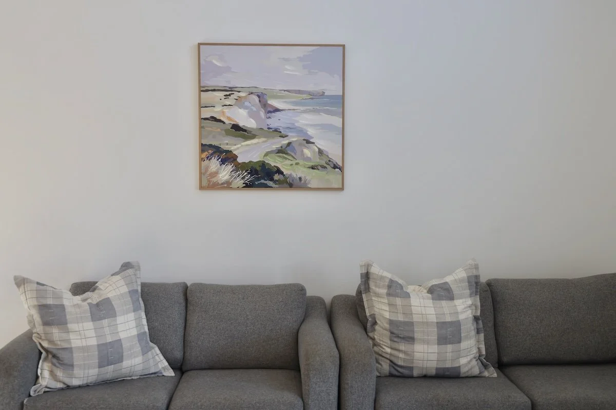 Living room with two gray sofas and plaid pillows, framed landscape painting of cliffs and ocean on wall.