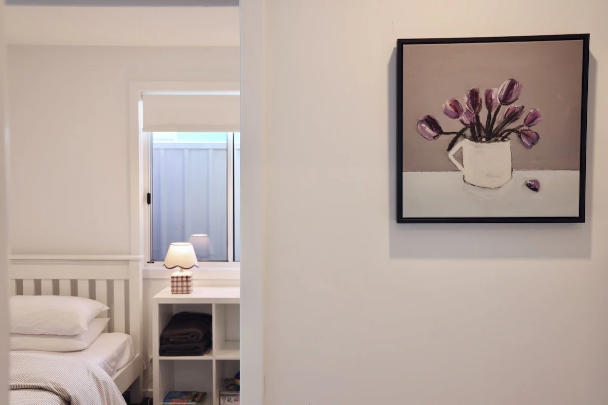 A bedroom with a white bed, a nightstand, a window, and a framed painting of a vase with purple tulips hanging on the wall.