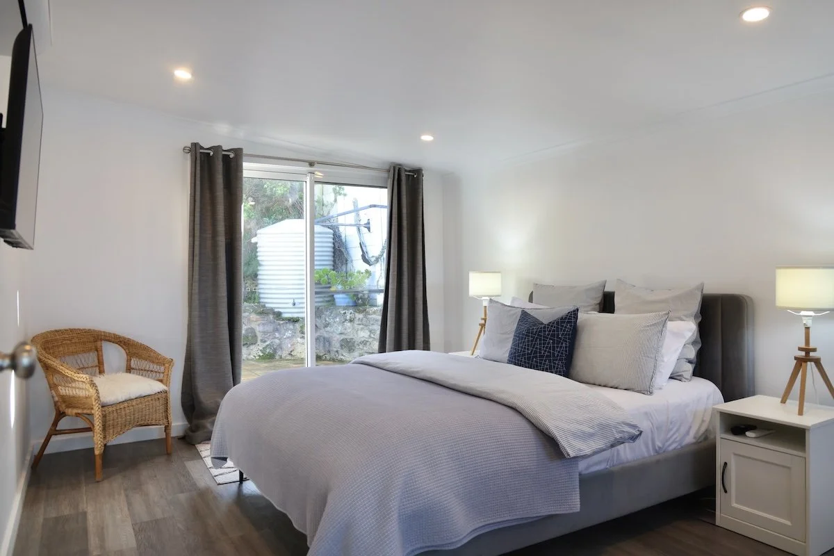 A cozy bedroom with a large bed, gray headboard, and multiple pillows, beside a white nightstand with a lamp. A small wicker chair with a cushion is in the corner, and a sliding glass door with curtains opens to an outdoor area with a water tank and 