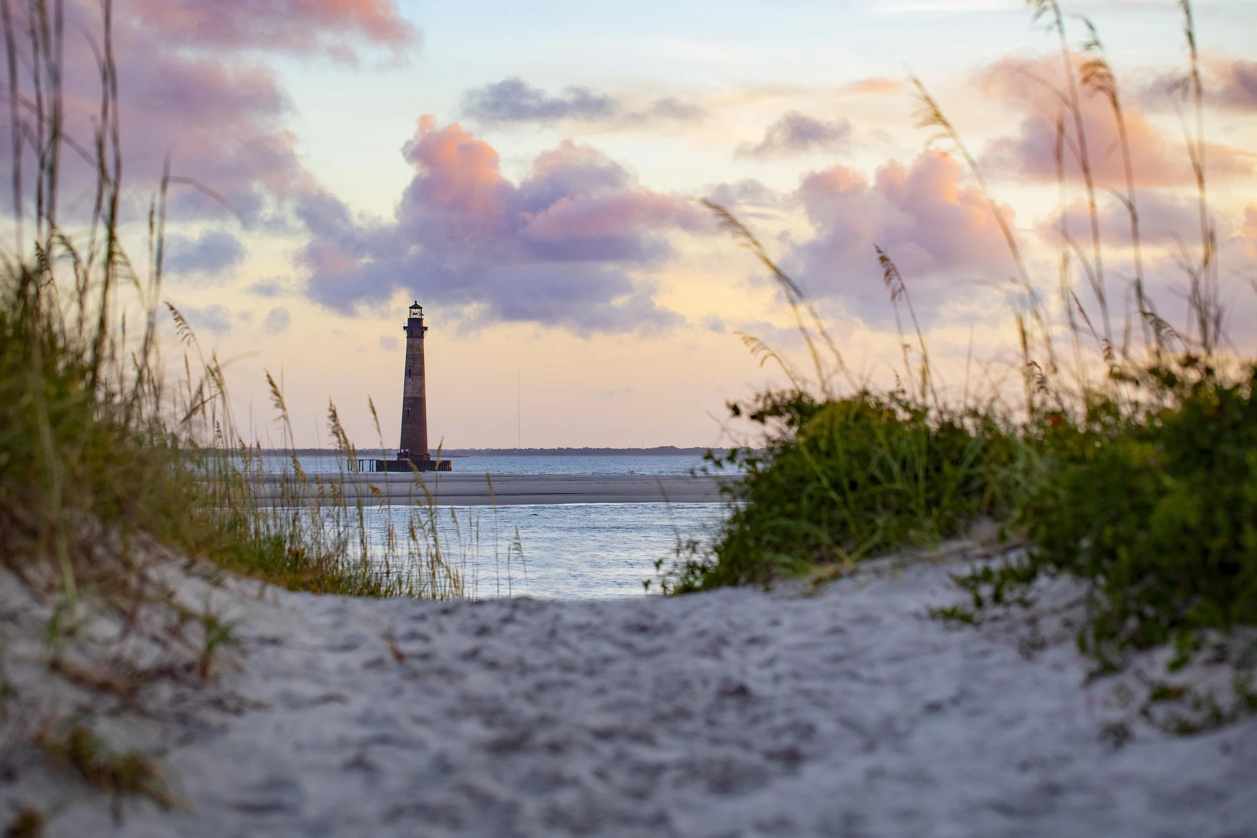 A lighthouse on a small island, viewed from a sandy beach with grass and shrubs, during sunset with colorful clouds in the sky.
