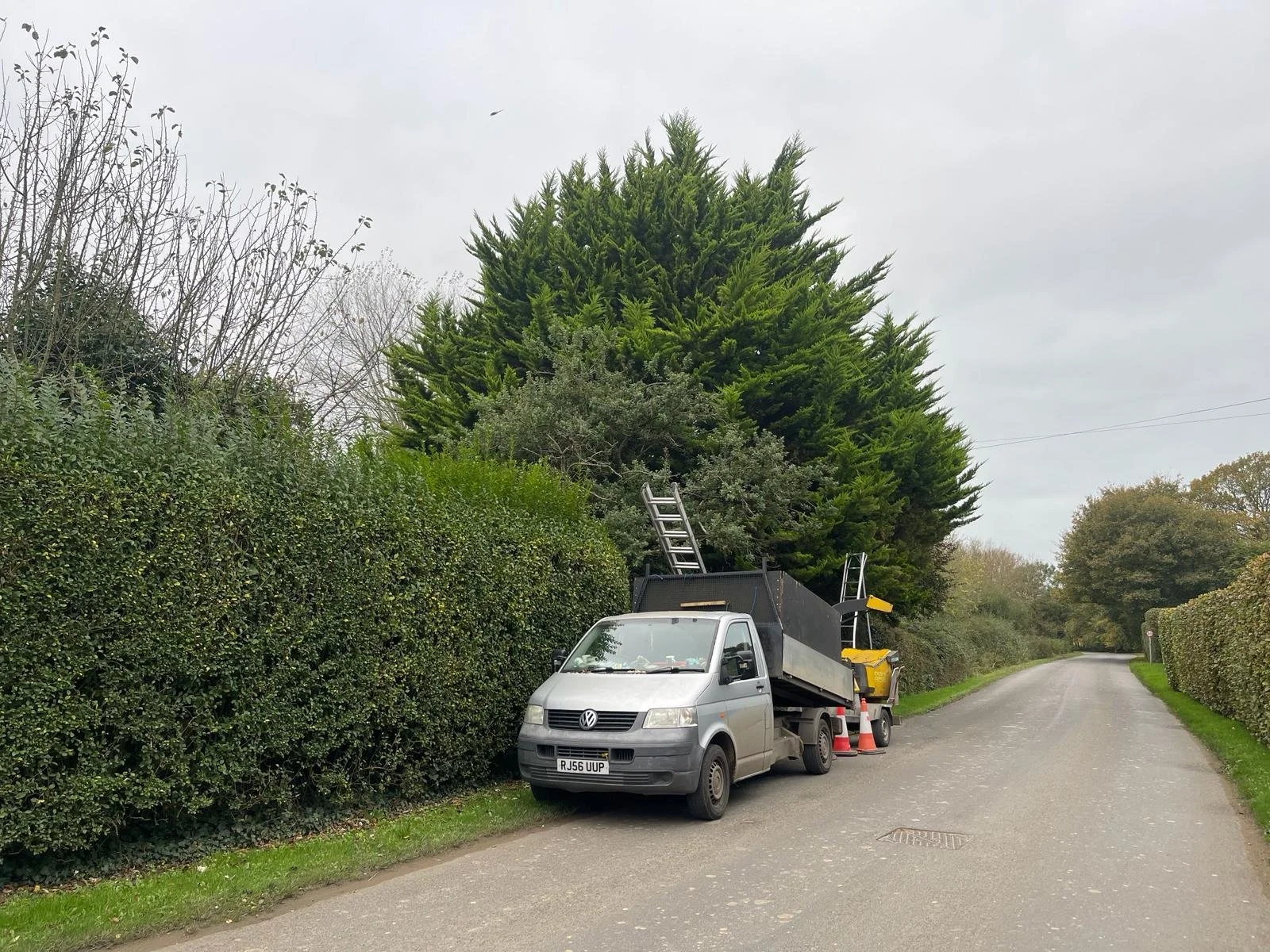 A construction or maintenance vehicle parked on the side of a rural road, with ladders and traffic cones nearby, surrounded by green hedges and trees.