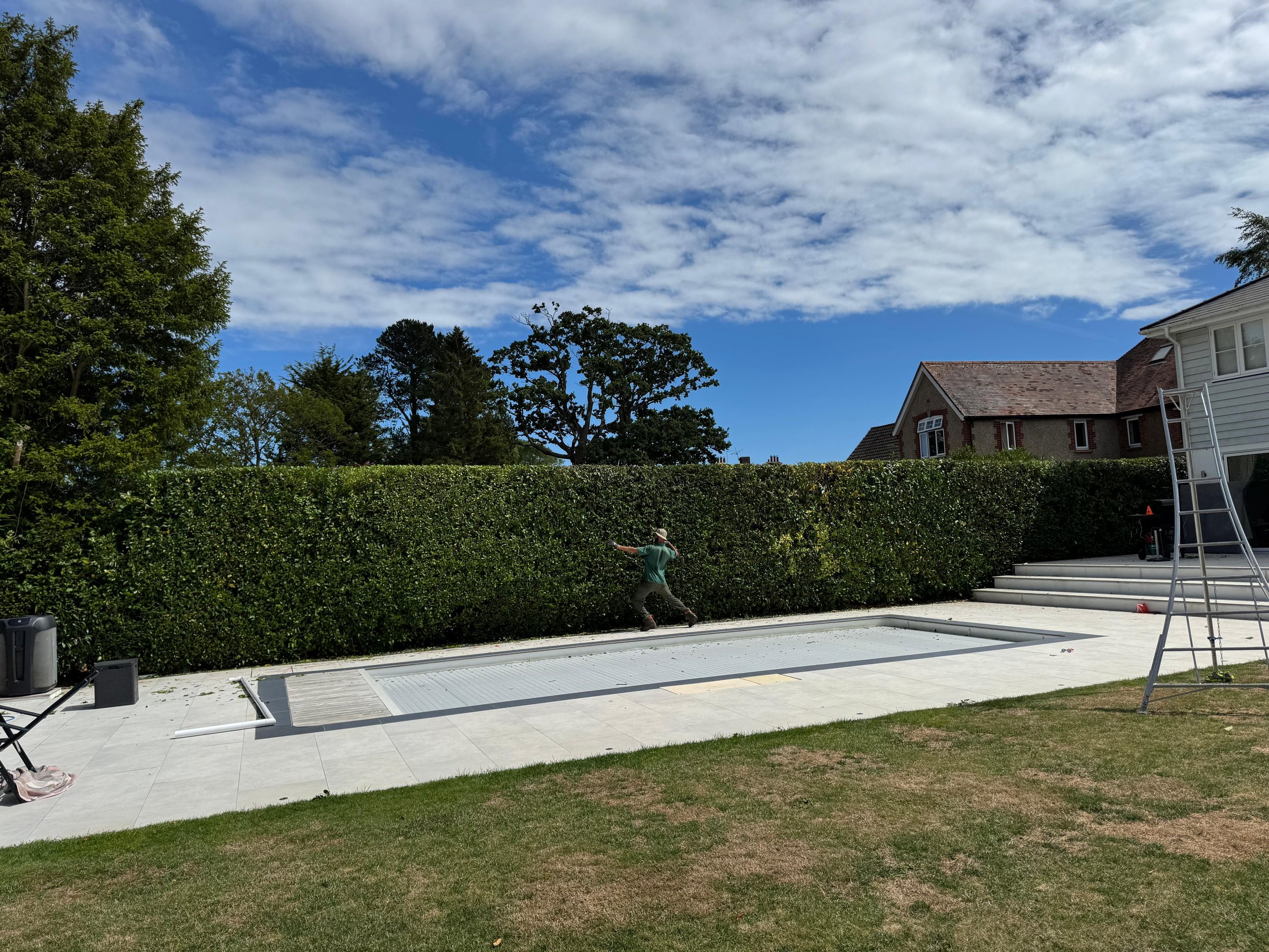 Person working on swimming pool cover in backyard with hedges, trees, house, and tools nearby.
