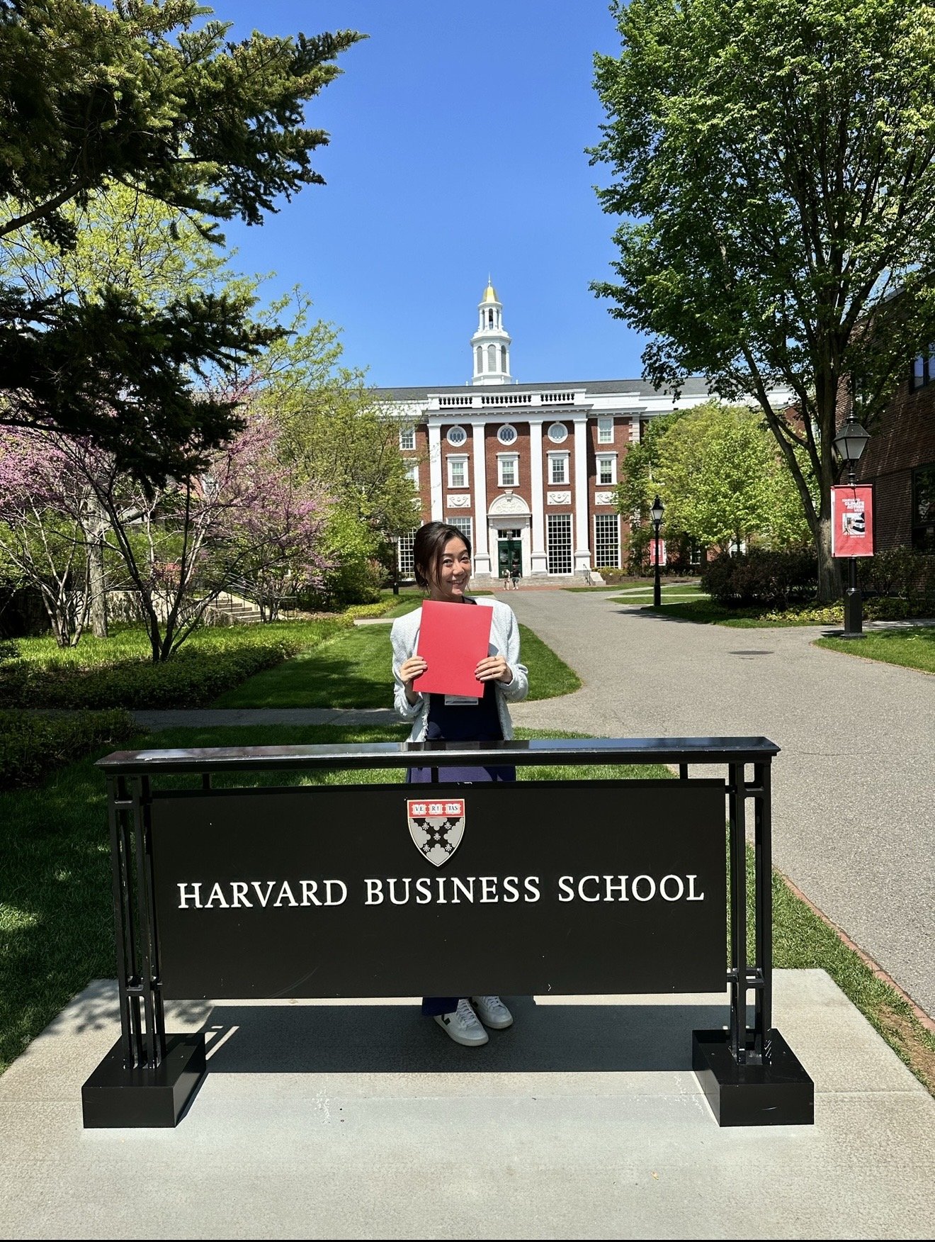 A woman standing behind a black Harvard Business School sign outdoors, holding a red folder, with a red brick building and trees in the background on a sunny day.