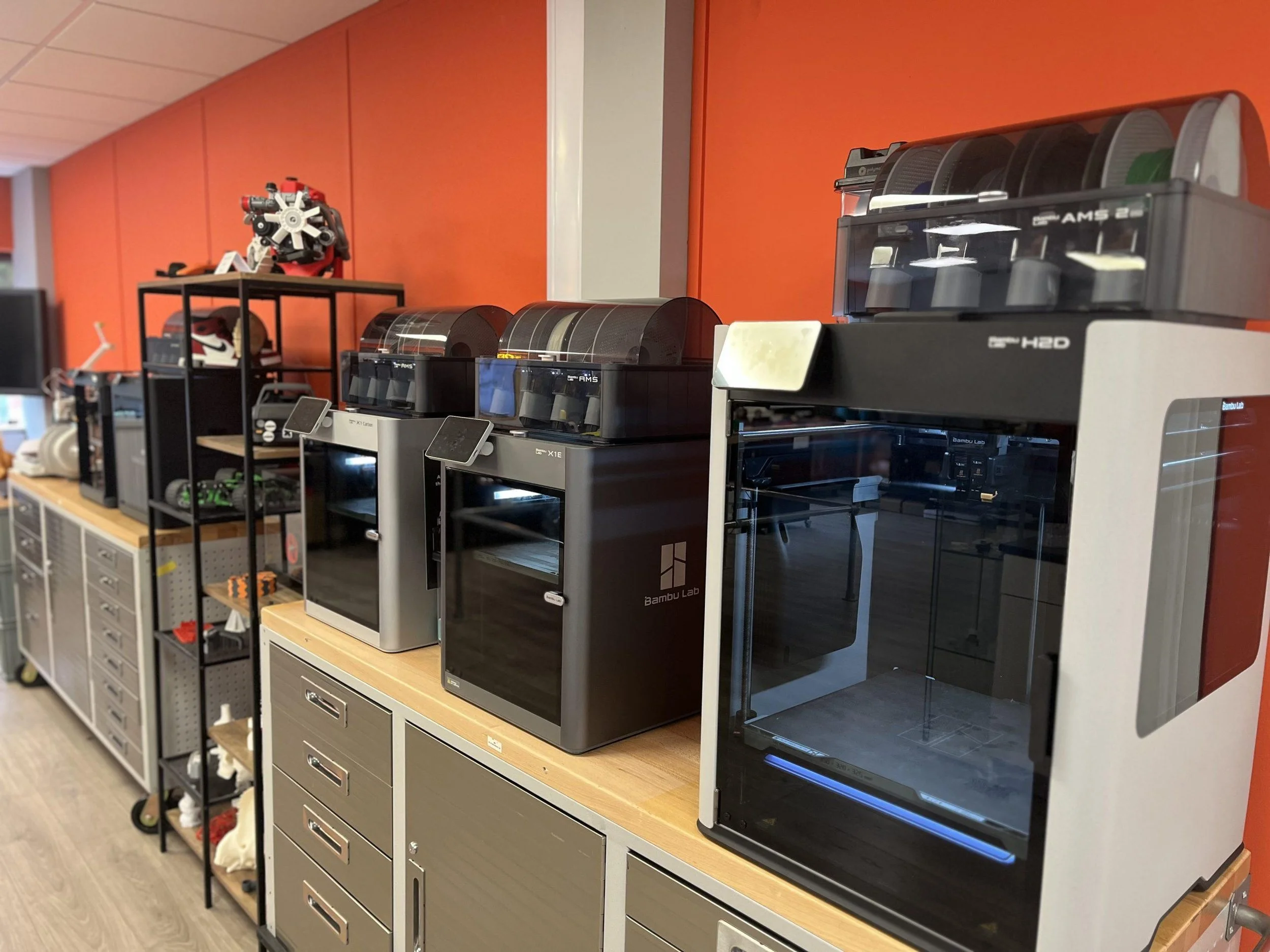 A row of 3D printers and filament spools on a table against an orange wall in a manufacturing or prototyping lab.