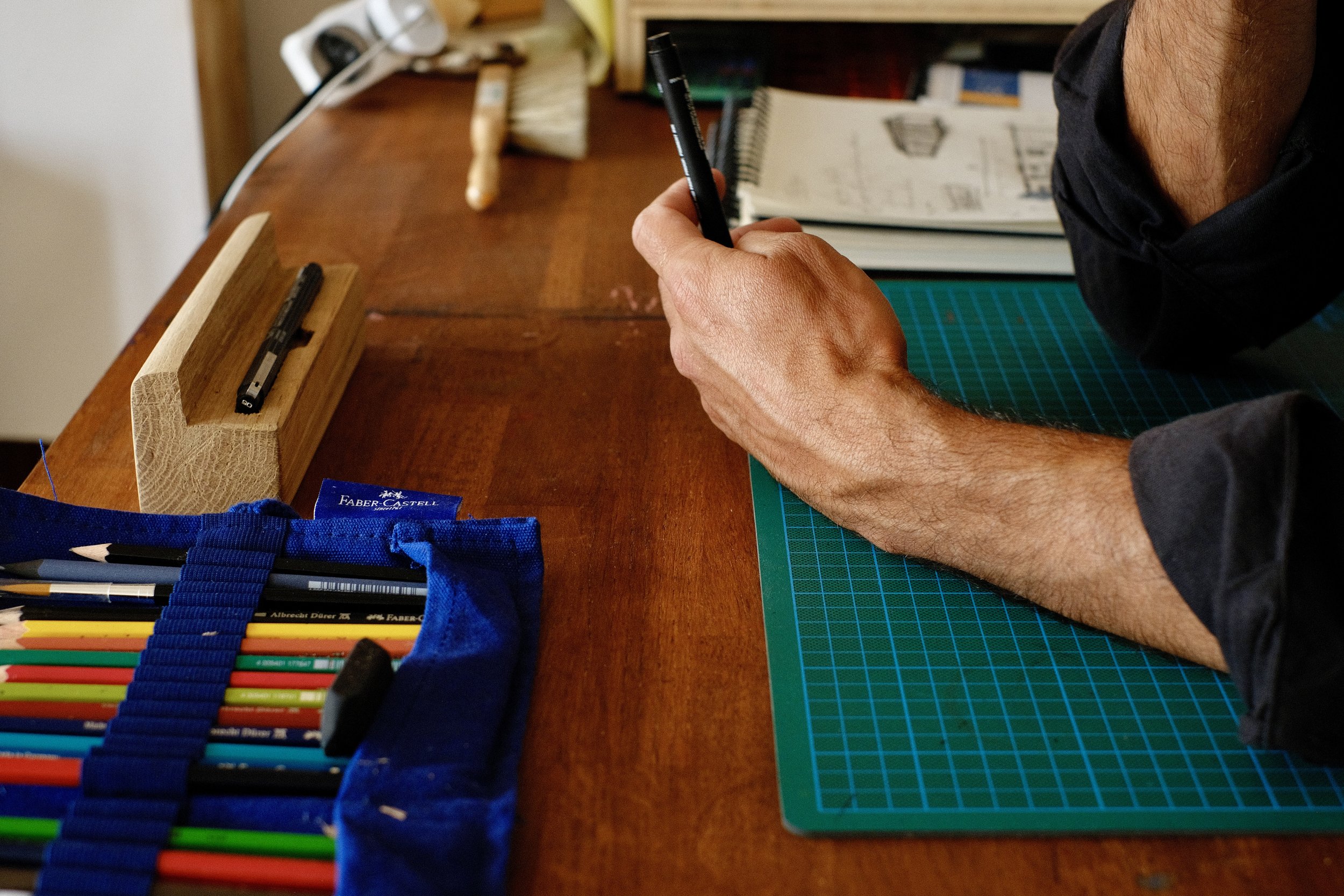 Une personne tient un marqueur noir dans sa main au-dessus d'un bureau en bois avec des crayons de couleur organisés dans une poche en tissu à gauche. Sur le bureau, il y a un tapis de coupe vert, un bloc-notes, des livres, et des papiers. En arrière-plan, on voit une étagère avec des livres et des objets, et un câble électrique.