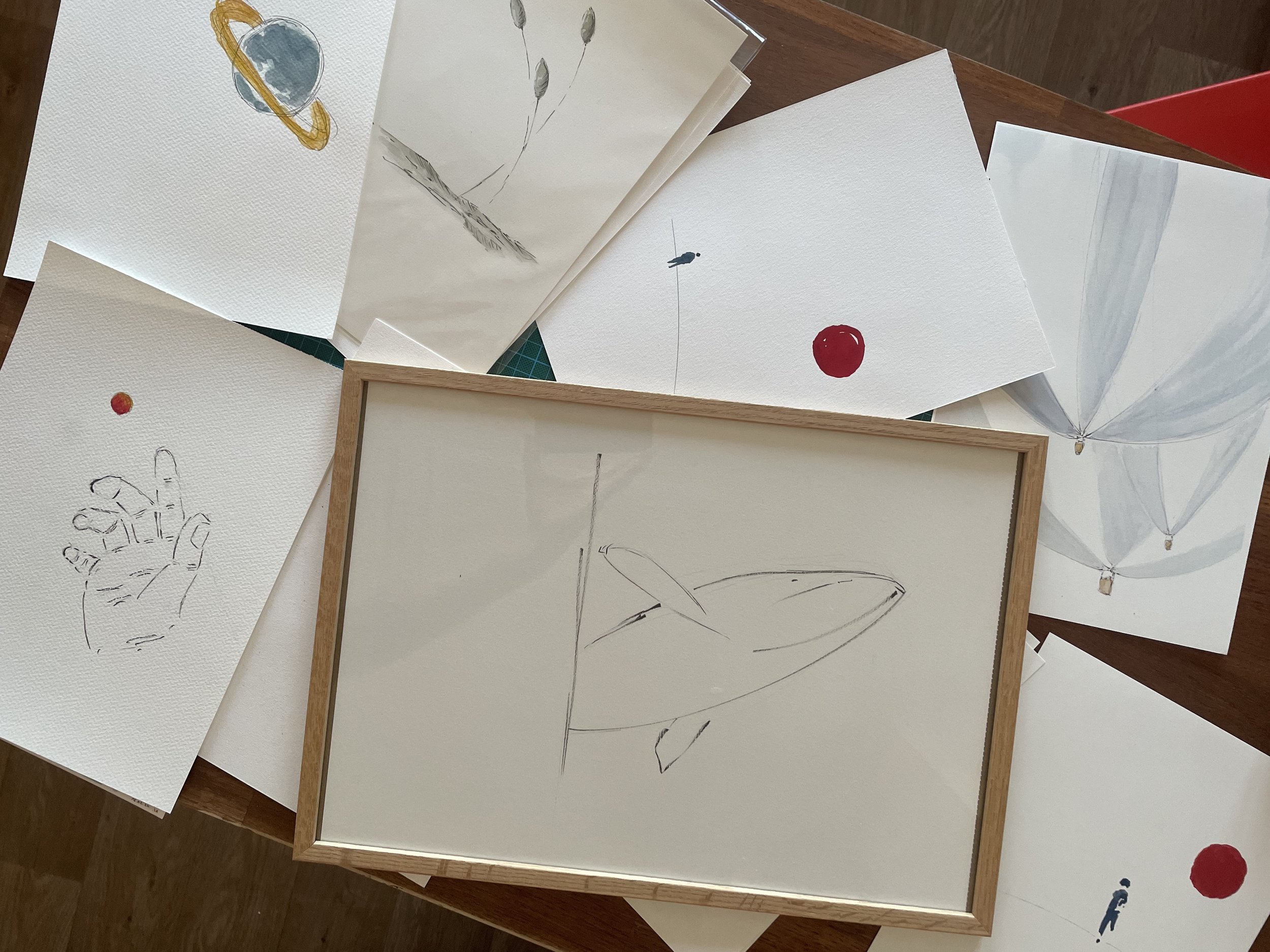 Plusieurs dessins faits à la main sur des feuilles de papier, dont un représentant une planète avec un anneau, des ballons, une main esquissée et un cercle rouge.