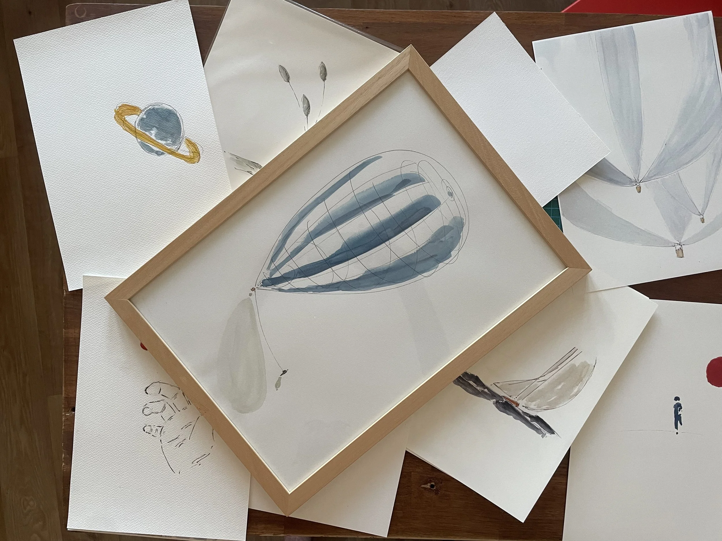 Collection de dessins et peintures d'objets flottants et de formes abstraites, encadrées ou sur papier, dont une représentation de Saturne, un ballon en forme d’œuf et un ballon volant dans un ciel clair.