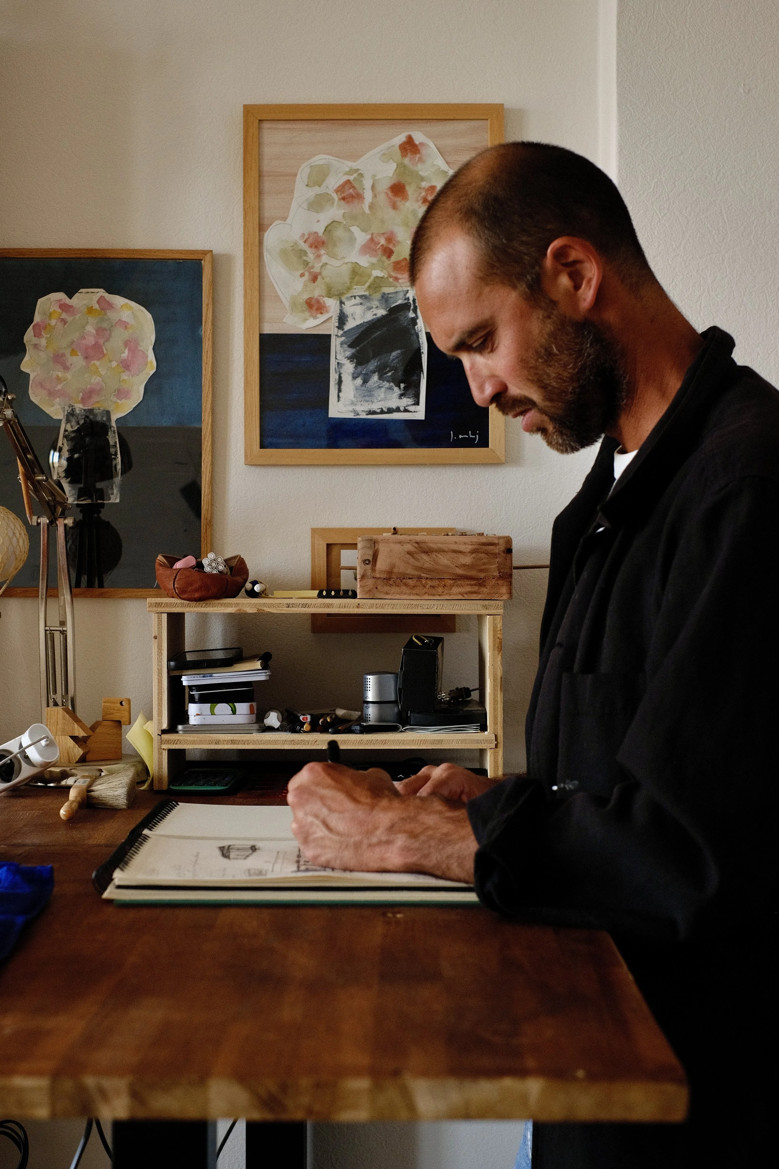 Un homme avec une barbe assis à une table en bois, concentré sur un dessin dans un carnet ouvert. En arrière-plan, des peintures abstraites colorées sont accrochées au mur, et une étagère en bois contient divers objets, dont un petit ordinateur, des accessoires, et des fournitures artistiques.