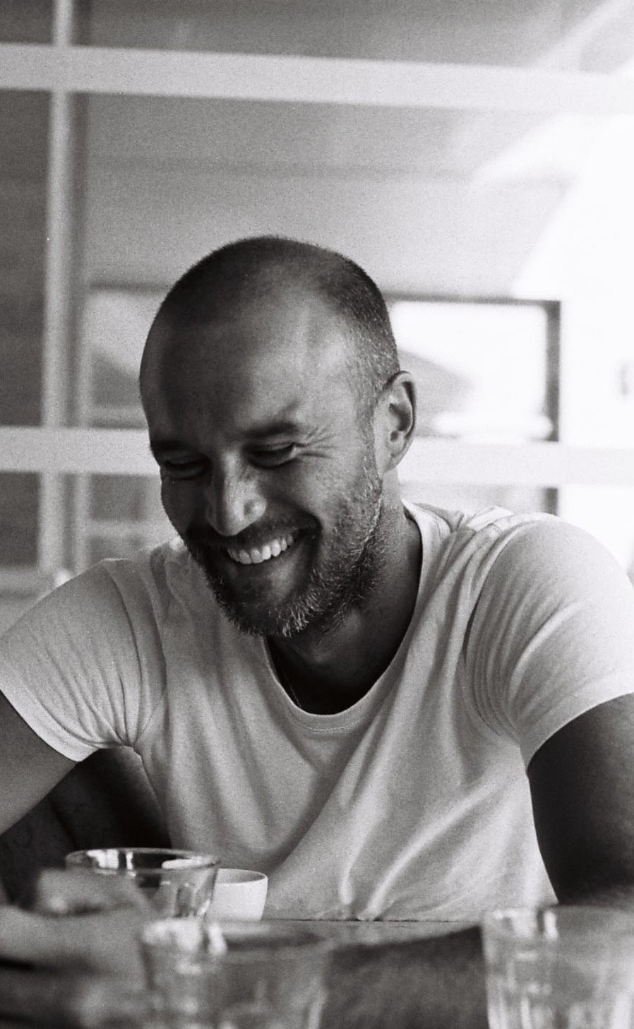 Un homme souriant en noir et blanc, portant un t-shirt blanc, assis à une table avec des verres transparent autour de lui.