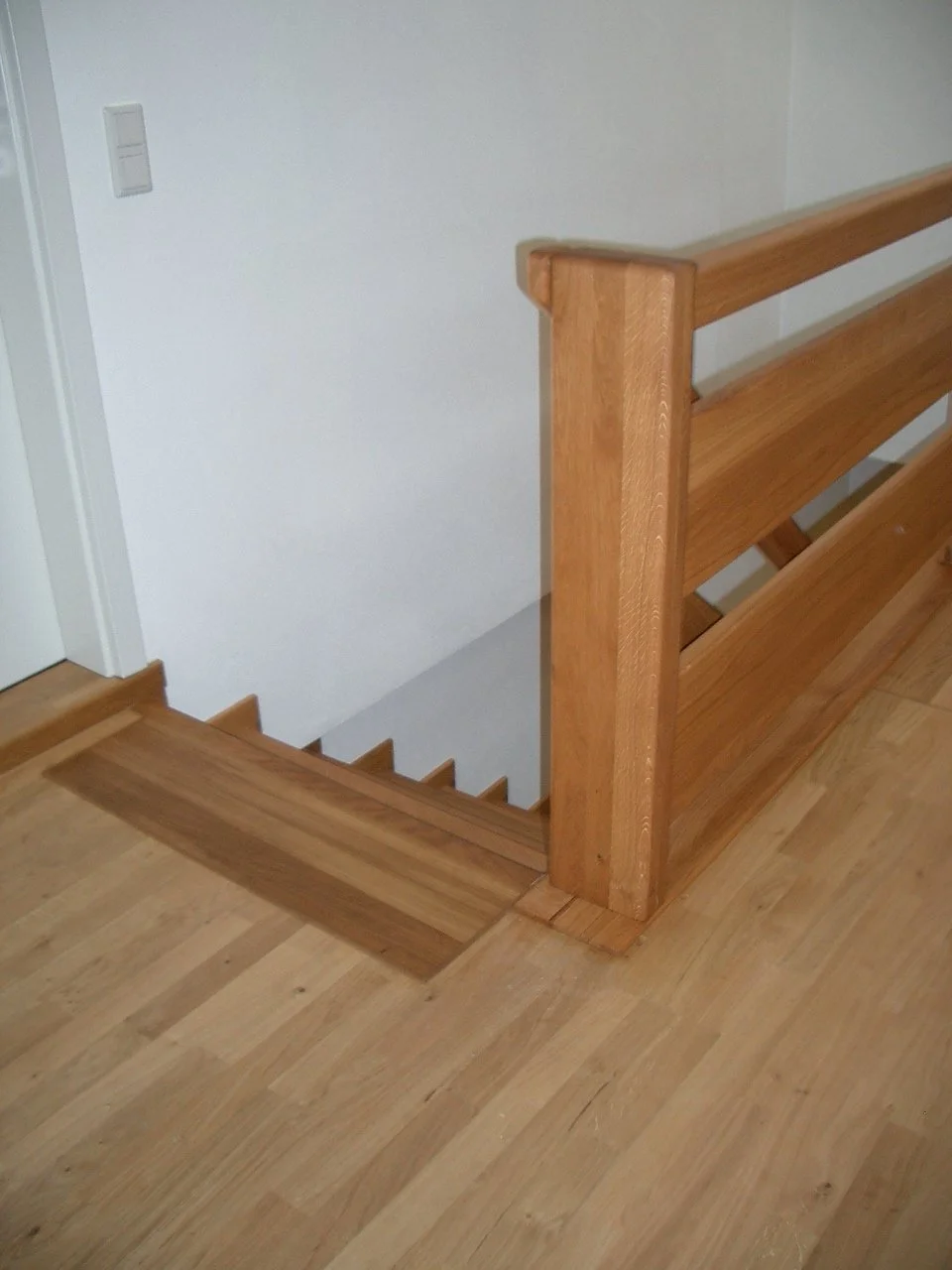 Wood staircase with a wooden railing leading down to a lower floor, next to a white wall and a light switch.