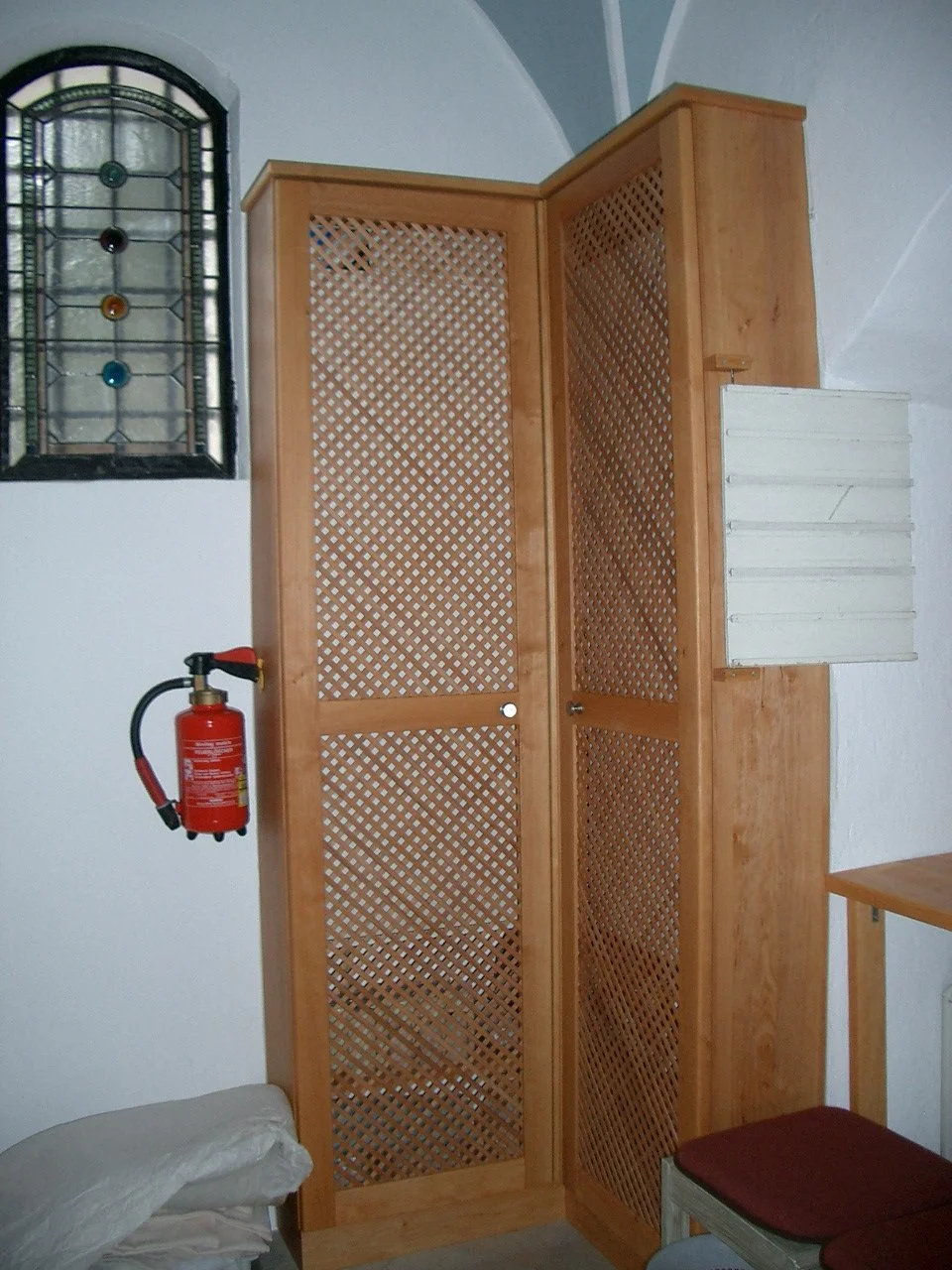A corner of a room with wooden woven cabinet doors, a small white shelf to the right, and a red fire extinguisher mounted on the wall. There is a cushion on a bench or stool in the lower right corner, and a decorative stained glass window on the left