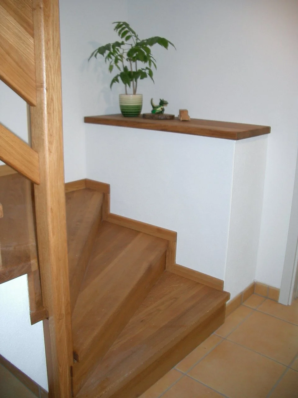 Wood staircase with a small landing, a white wall, and a wooden shelf with a potted plant and some decorative items.