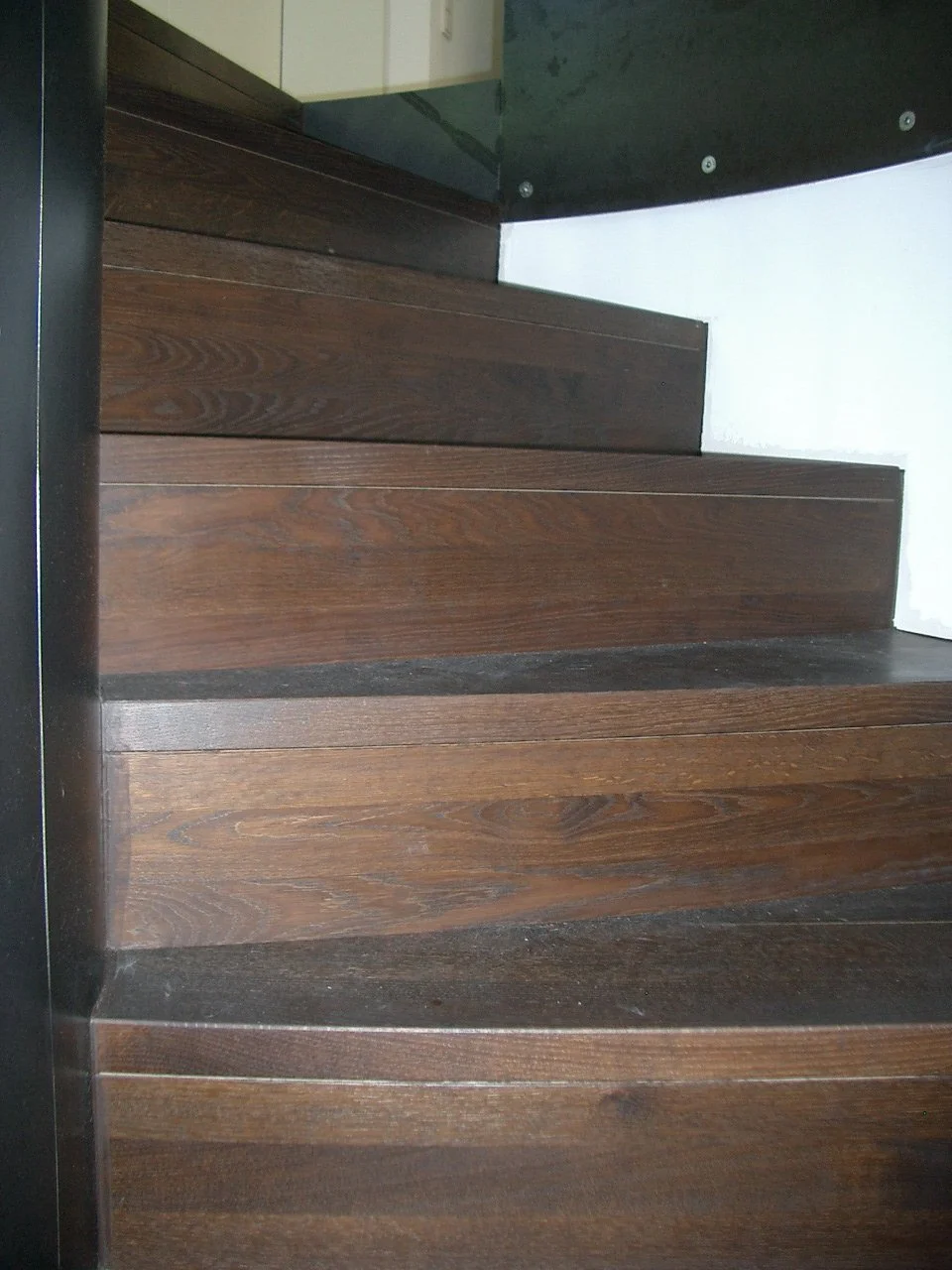 Wooden staircase with dark brown steps and black side frame, next to a white wall and black structural element.