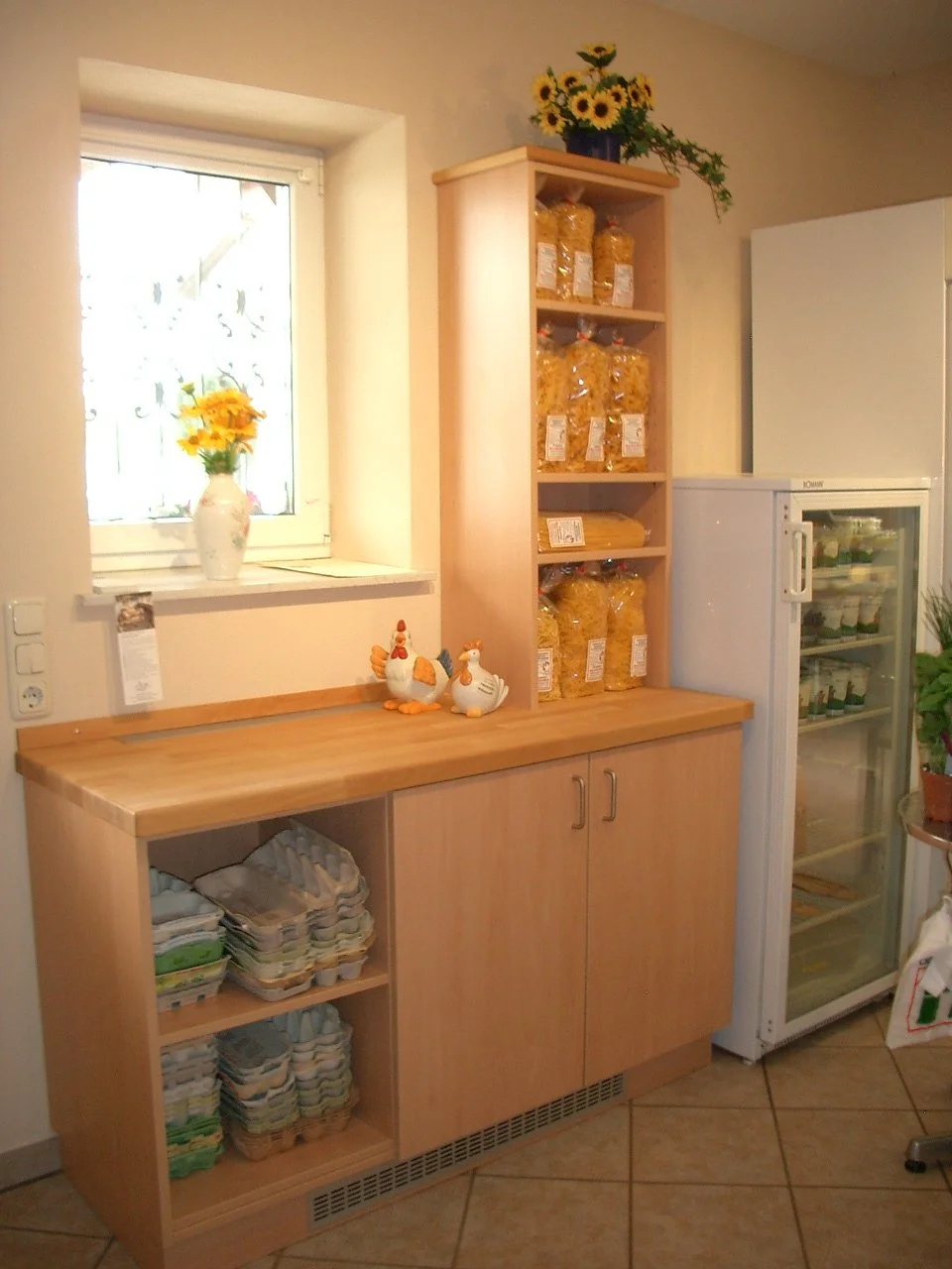 Wooden kitchen cabinet with a countertop, standing next to a small refrigerator and a window with a floral curtain. The cabinet has closed doors below and open shelves above filled with packaged pasta. On the countertop, there are ceramic chicken and