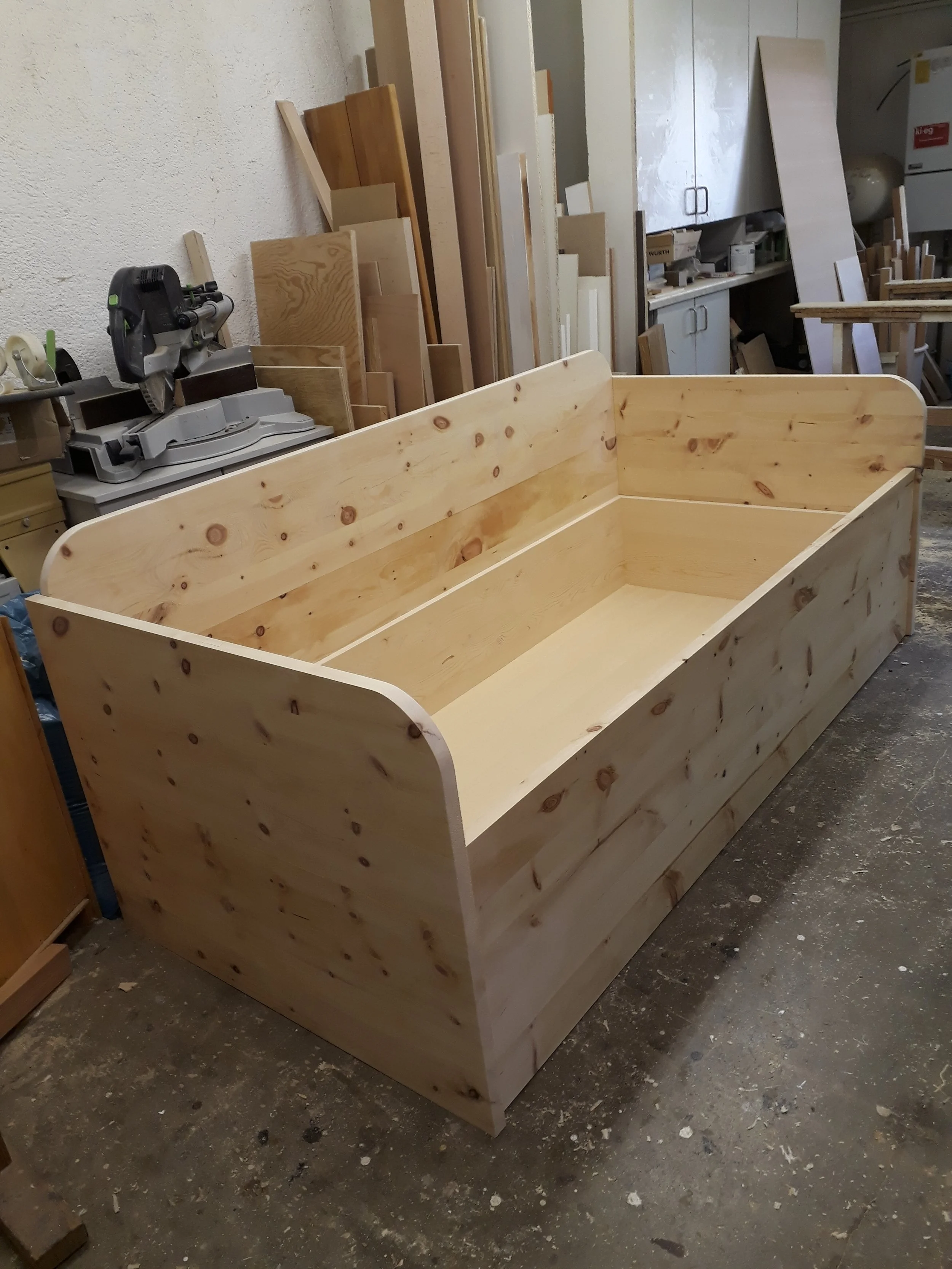 An unfinished wooden bed frame in a woodworking shop, with various wooden planks and tools in the background.