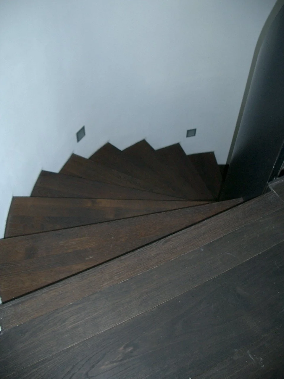 Top view of dark wooden staircase winding downward along a white wall.