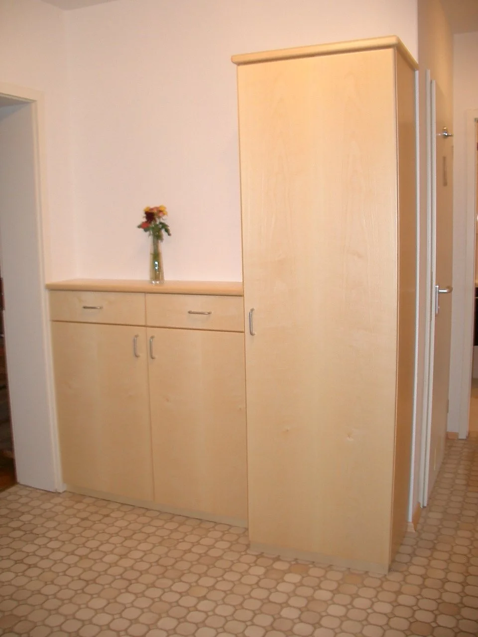 A room with a tall wooden cabinet, a shorter matching cabinet with a vase of flowers on top, and hexagonal tile flooring.