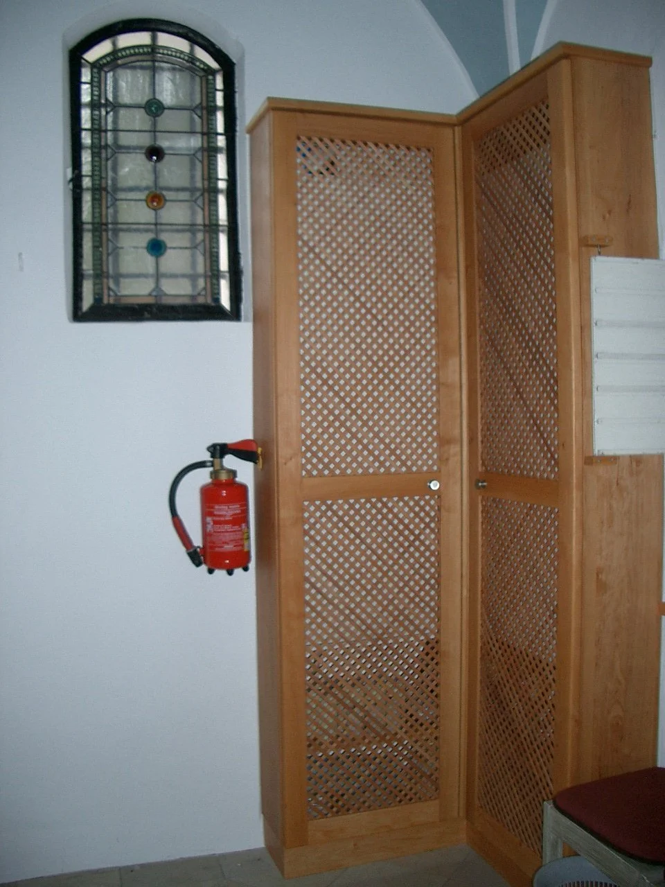 Wooden lattice storage cabinet next to a wall with a stained glass window and a mounted fire extinguisher.