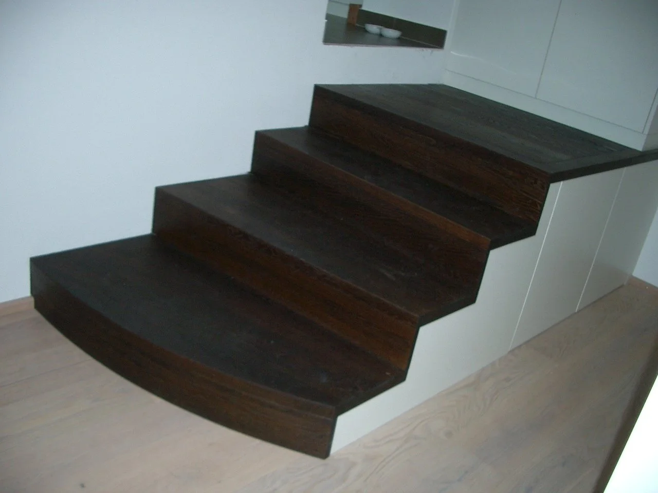 Interior of a home with a set of dark wooden stairs leading up to a white wall and cabinetry. The stairs have a curved bottom step and a small cutout in the wall near the top.