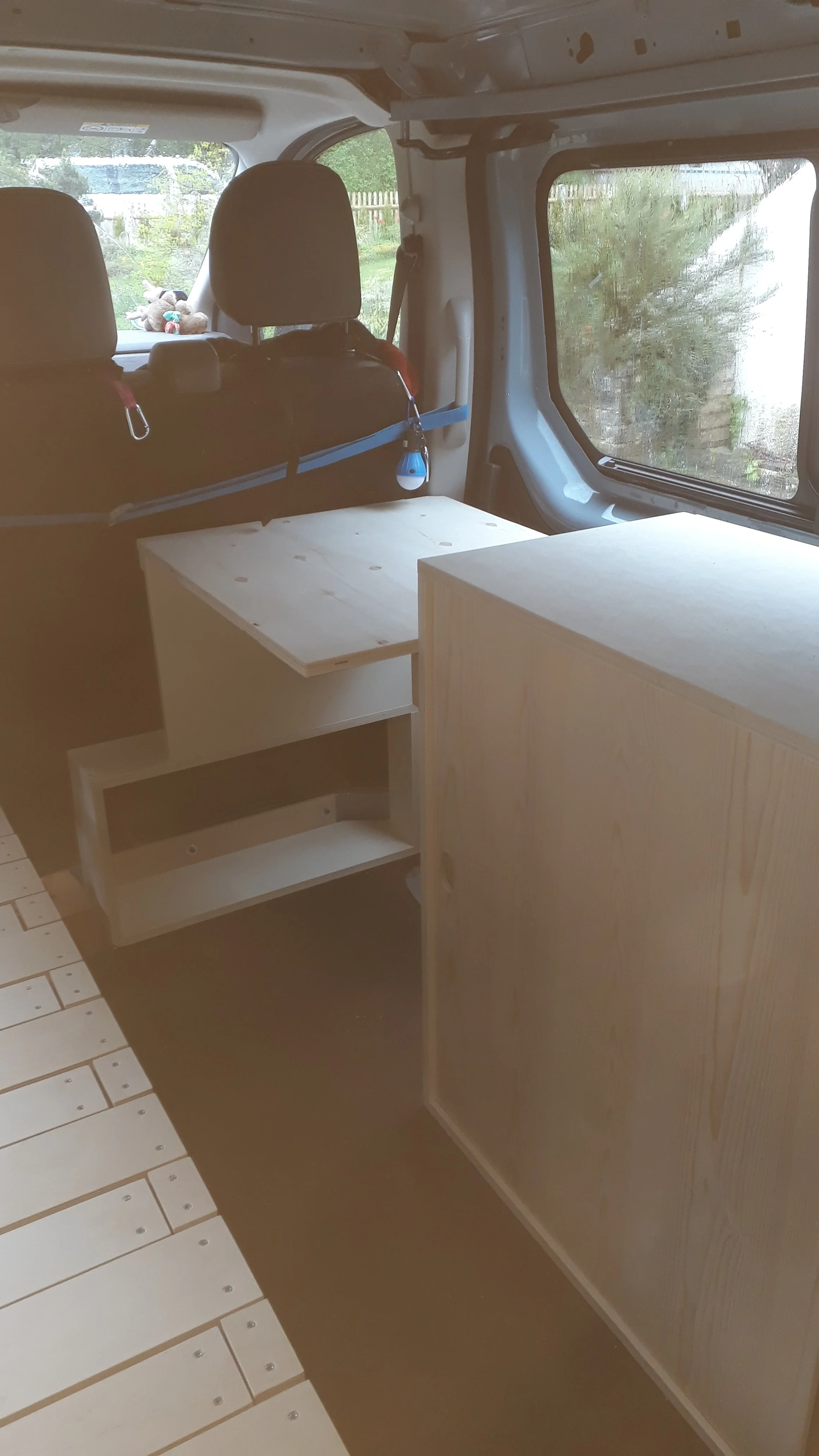 Interior of a camper van with unfinished wooden furniture, a window showing greenery outside, and a small wooden storage unit.