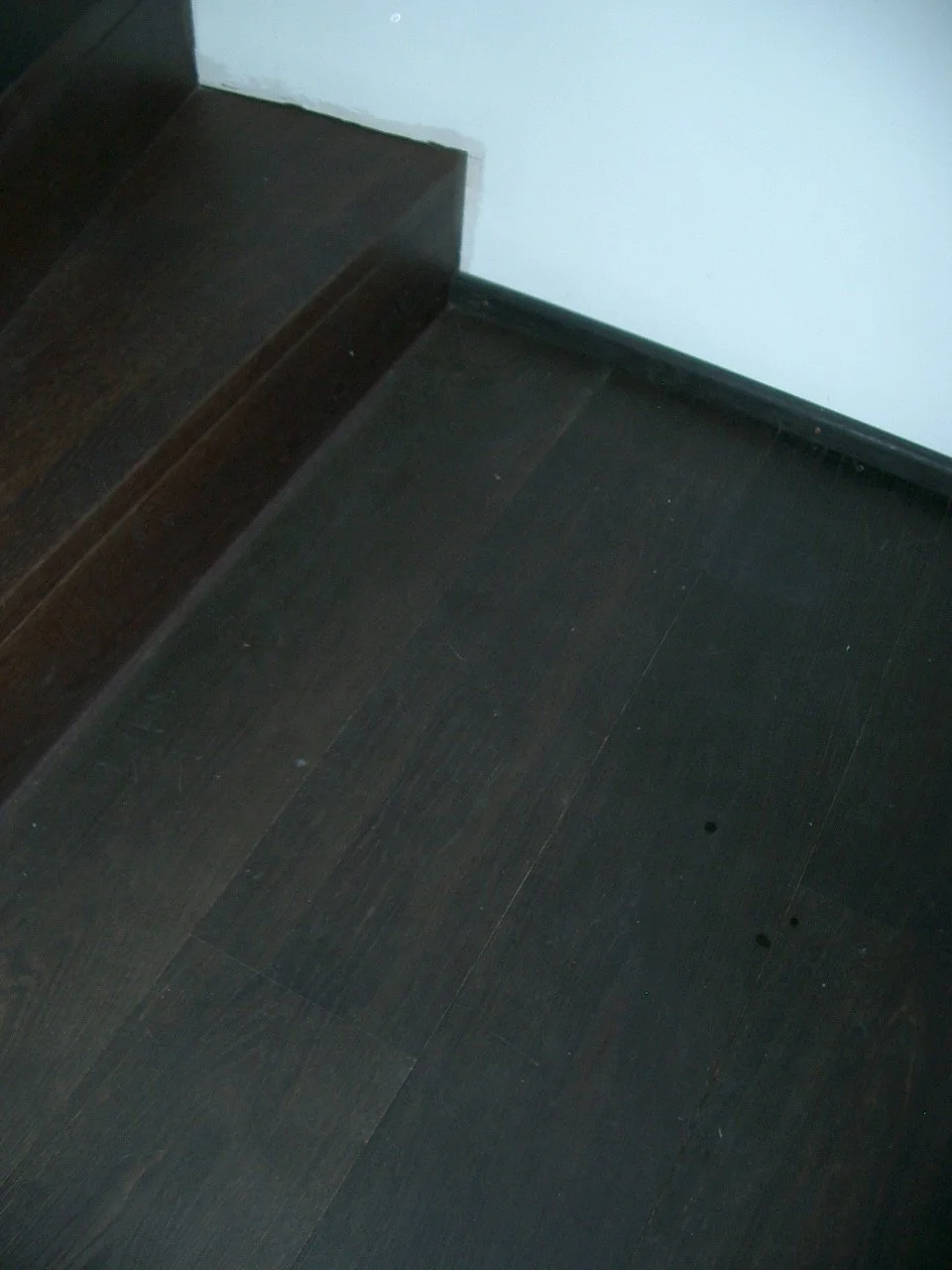 Close-up photo of dark wooden floor and a portion of a dark wooden stair with a white wall in the background.