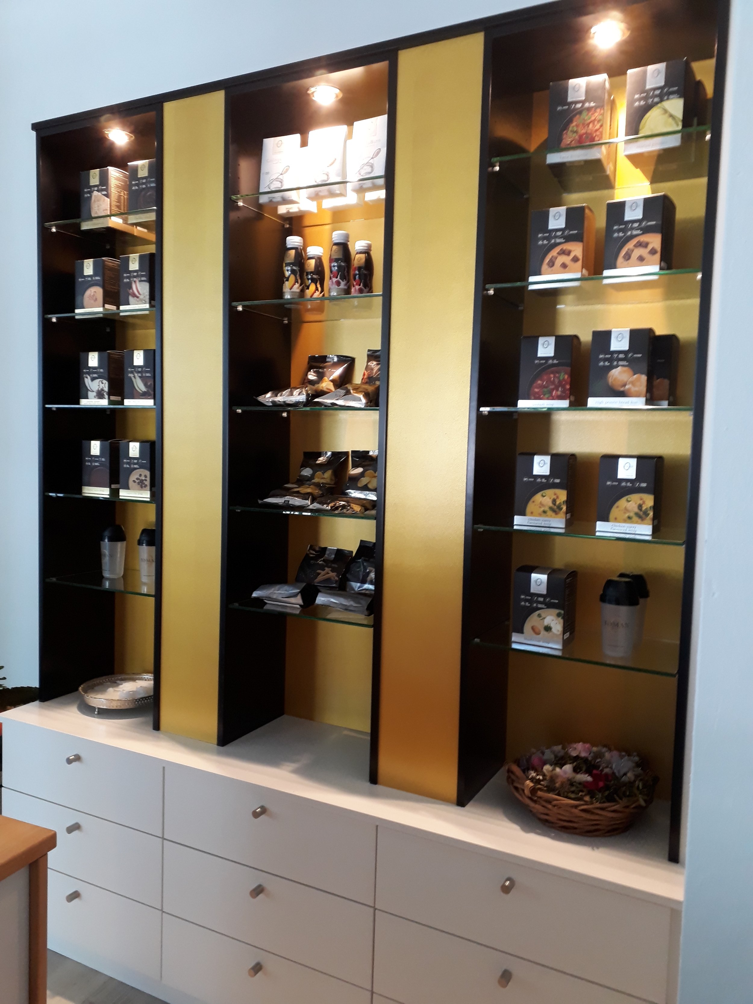 A white cabinet with drawers at the bottom and a black and yellow decorative wall shelf above. The shelf contains various packaged food items, bottles, and a woven basket with flowers on the right.