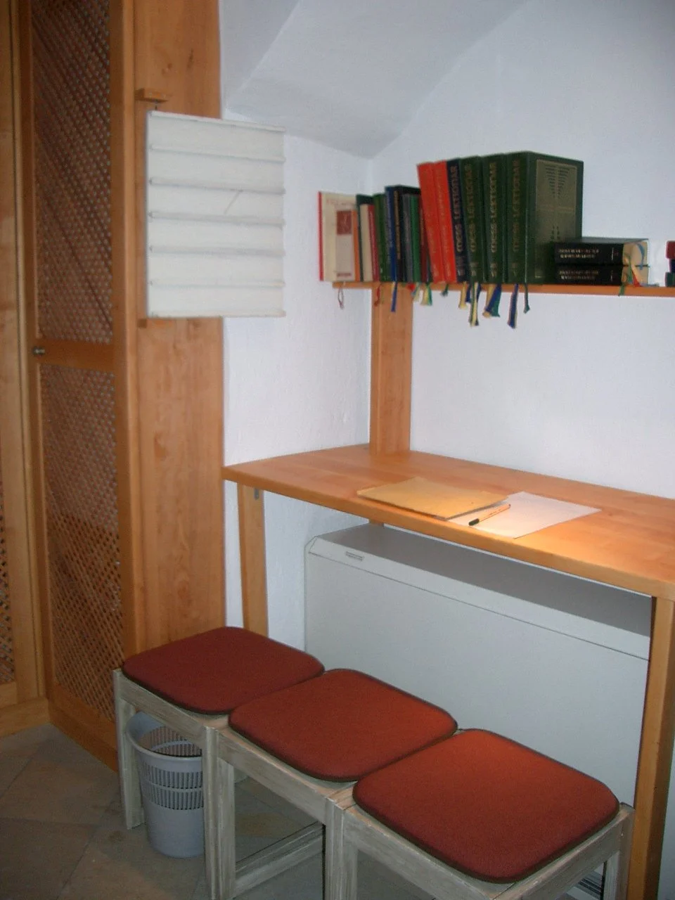 A small bookshelf with books, a desk with papers and a pen, and three red-cushioned stools underneath, in a cozy corner of a room.