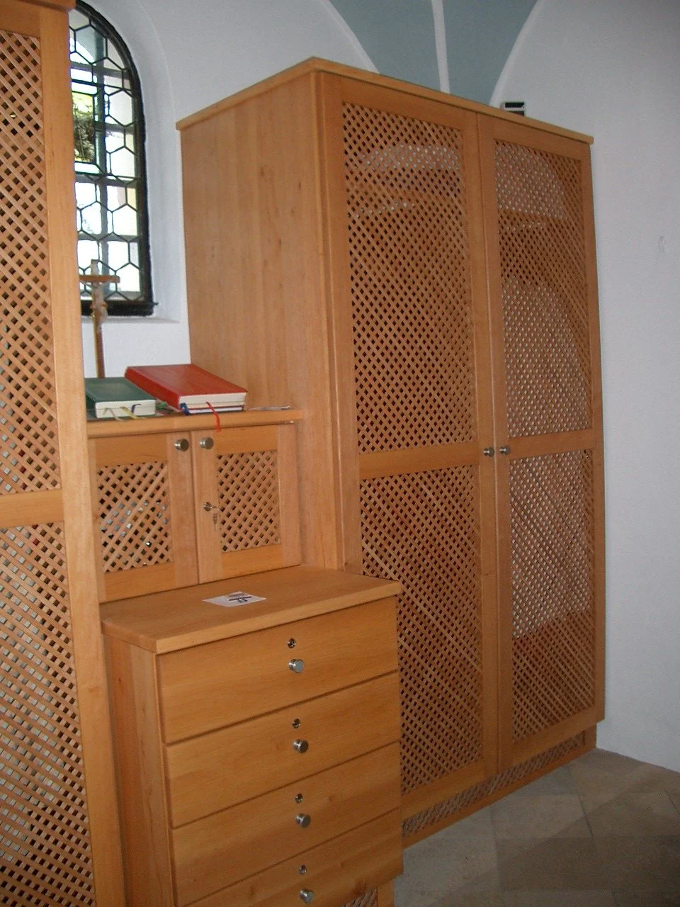 Interior view of a wooden cabinet with lattice doors and a small chest of drawers, located near a stained glass window in a room with arched ceiling.
