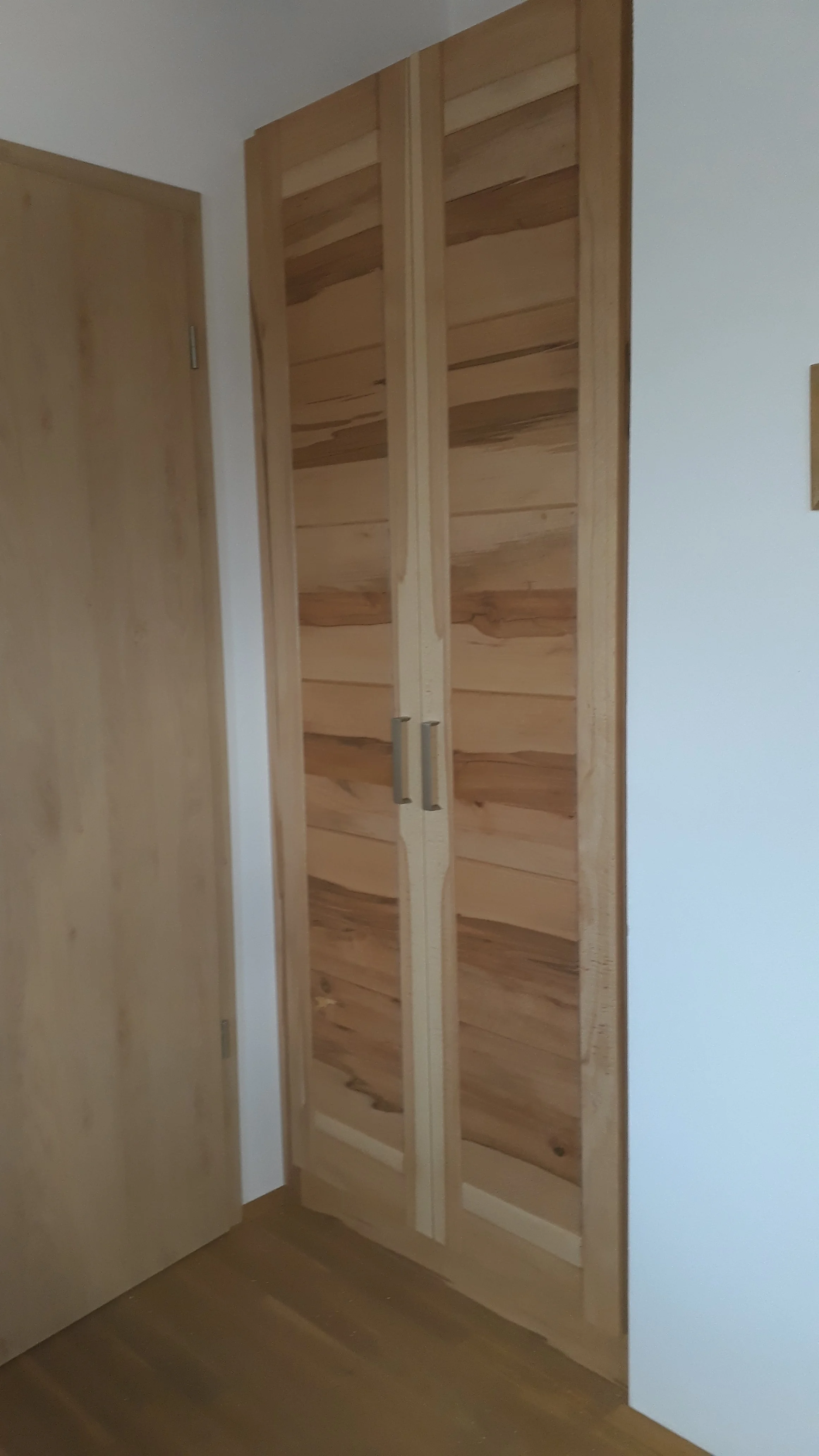 Interior view of a wooden wardrobe with two doors, situated next to a light-colored wall and adjacent to a wooden door.