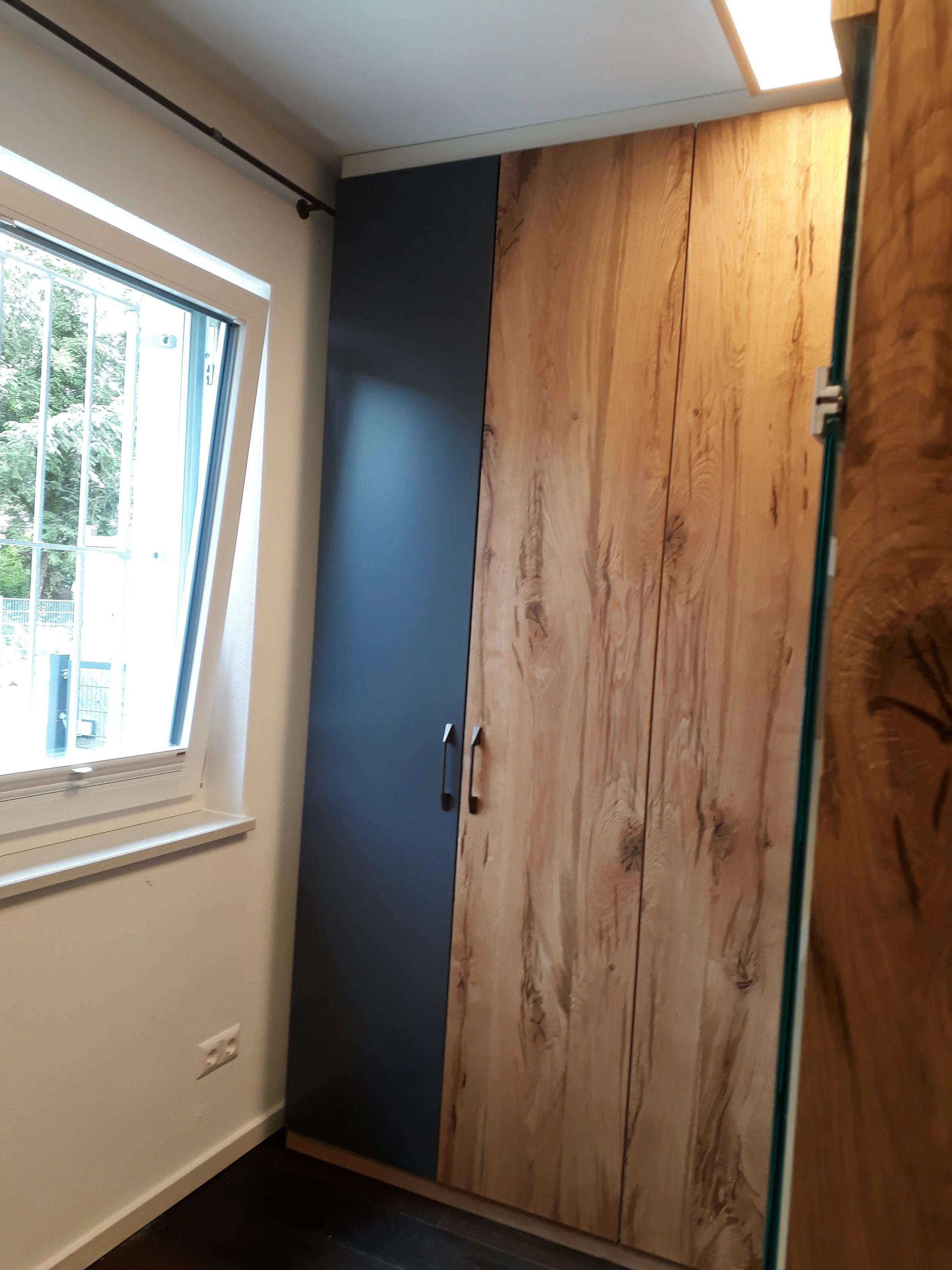 Close-up of a modern cabinet with a black door and wood grain finish, placed under a window in a room with white walls and a dark floor.