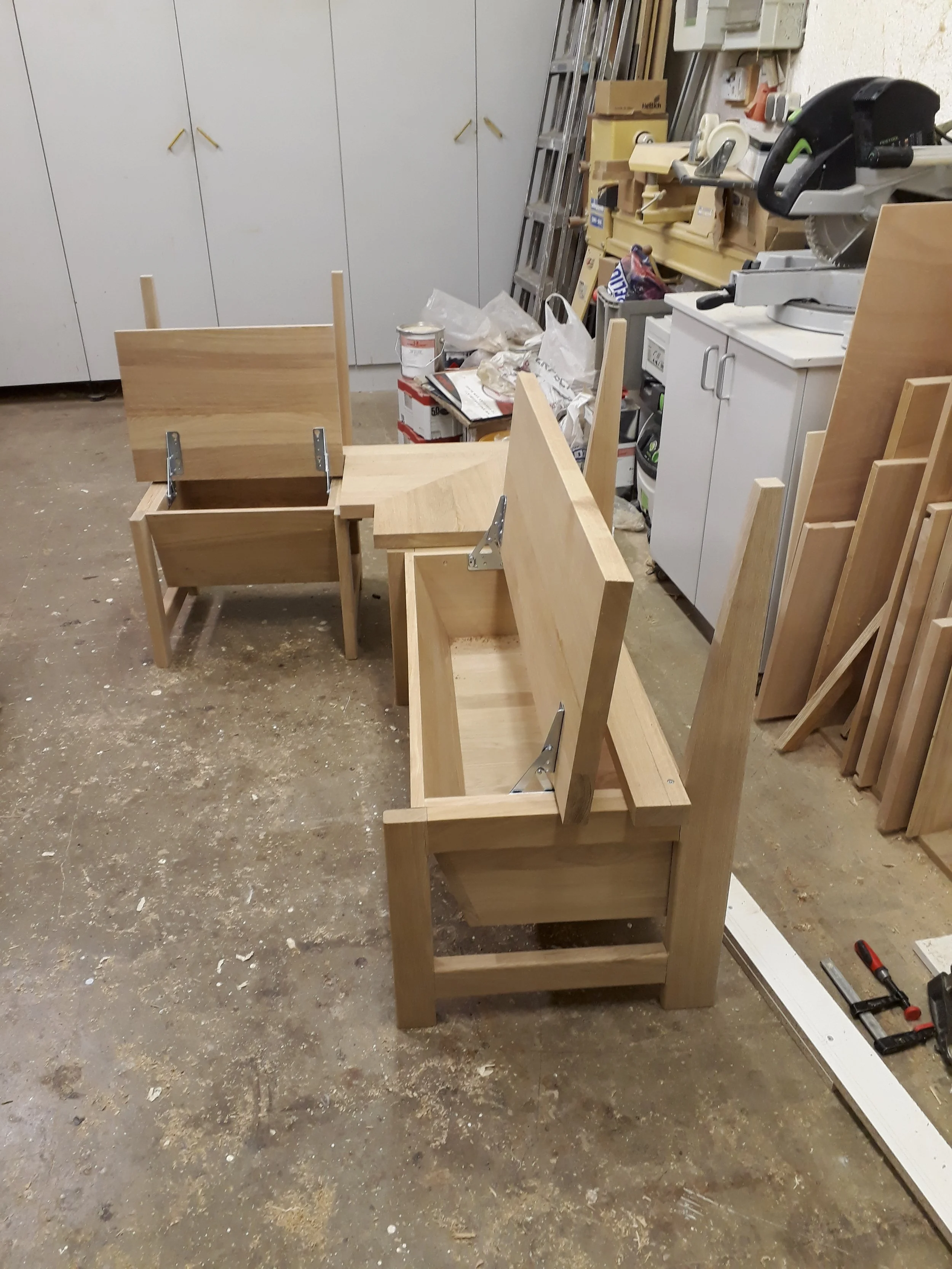 Unfinished wooden furniture in a workshop, with a partially assembled wooden bench visible in the foreground and tools, supplies, and a ladder in the background.